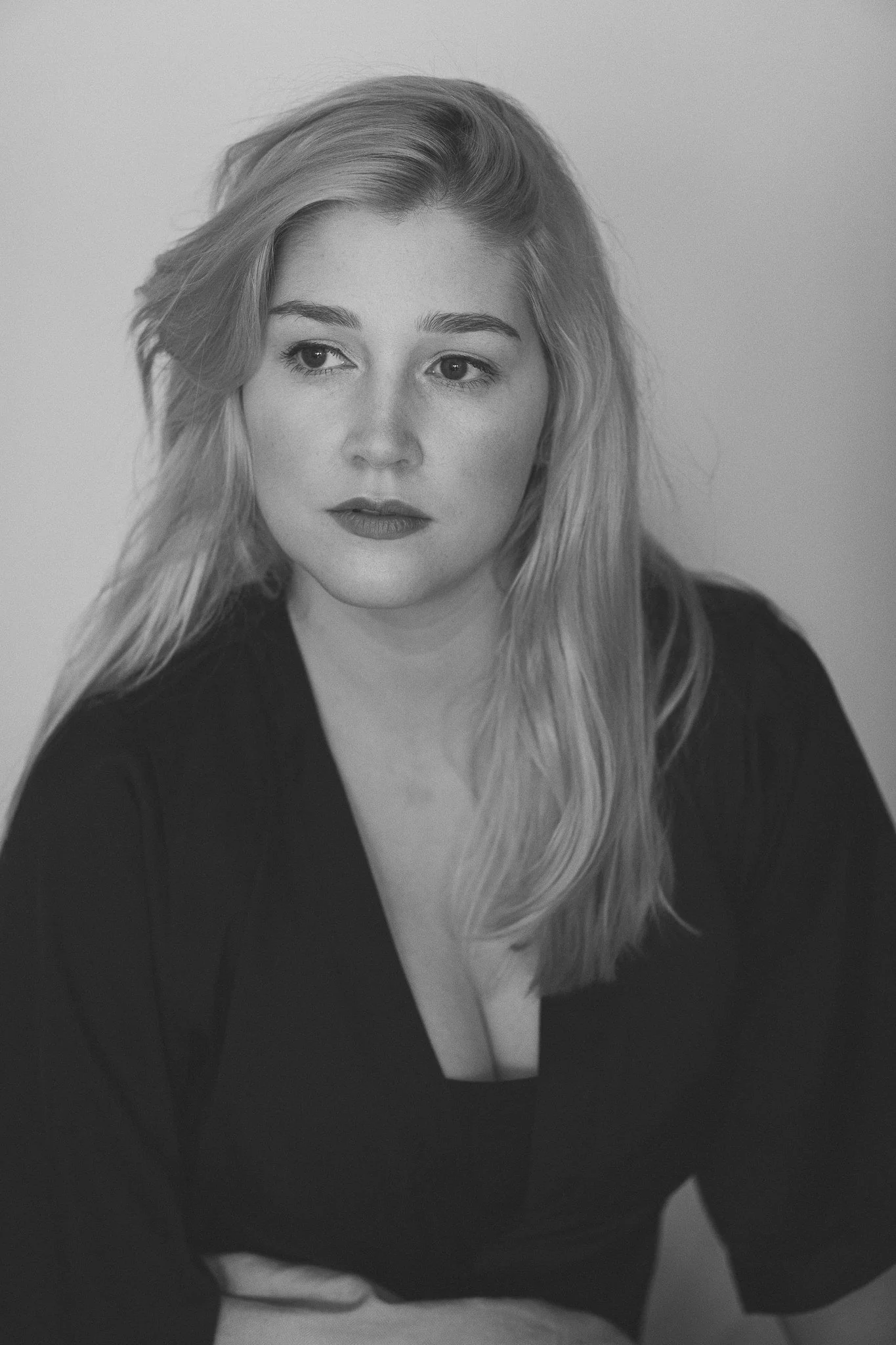 Schwarz-weiße Porträtaufnahme einer jungen Frau mit langem, lockigem Haar, die nach links schaut, trägt dunkel farbige Kleidung, vor hellem Hintergrund.