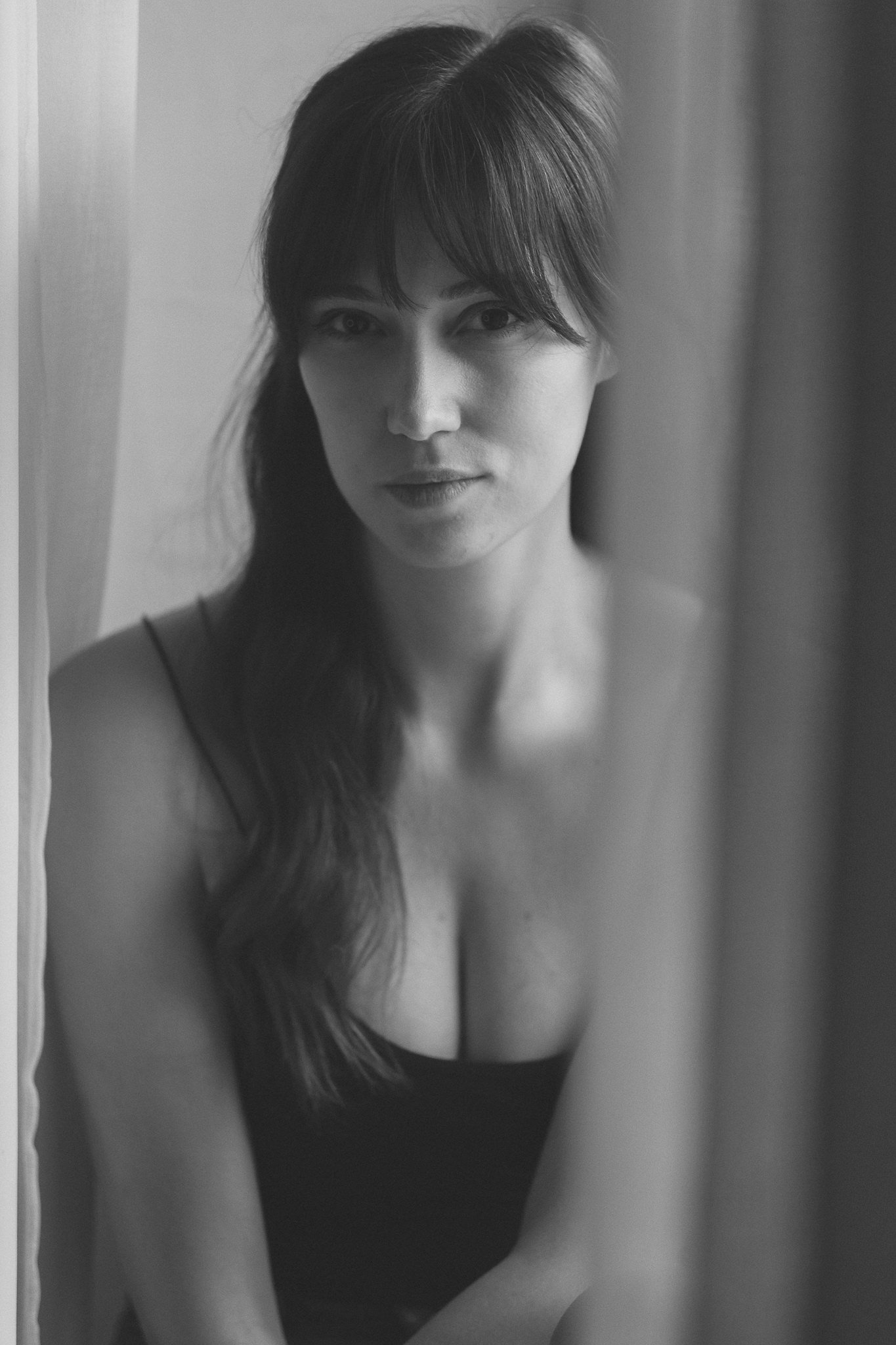 Schwarz-Weiß-Fotografie einer jungen Frau mit dunklen Haaren und natürlichem Gesichtsausdruck, die vor einem Vorhang posiert.
