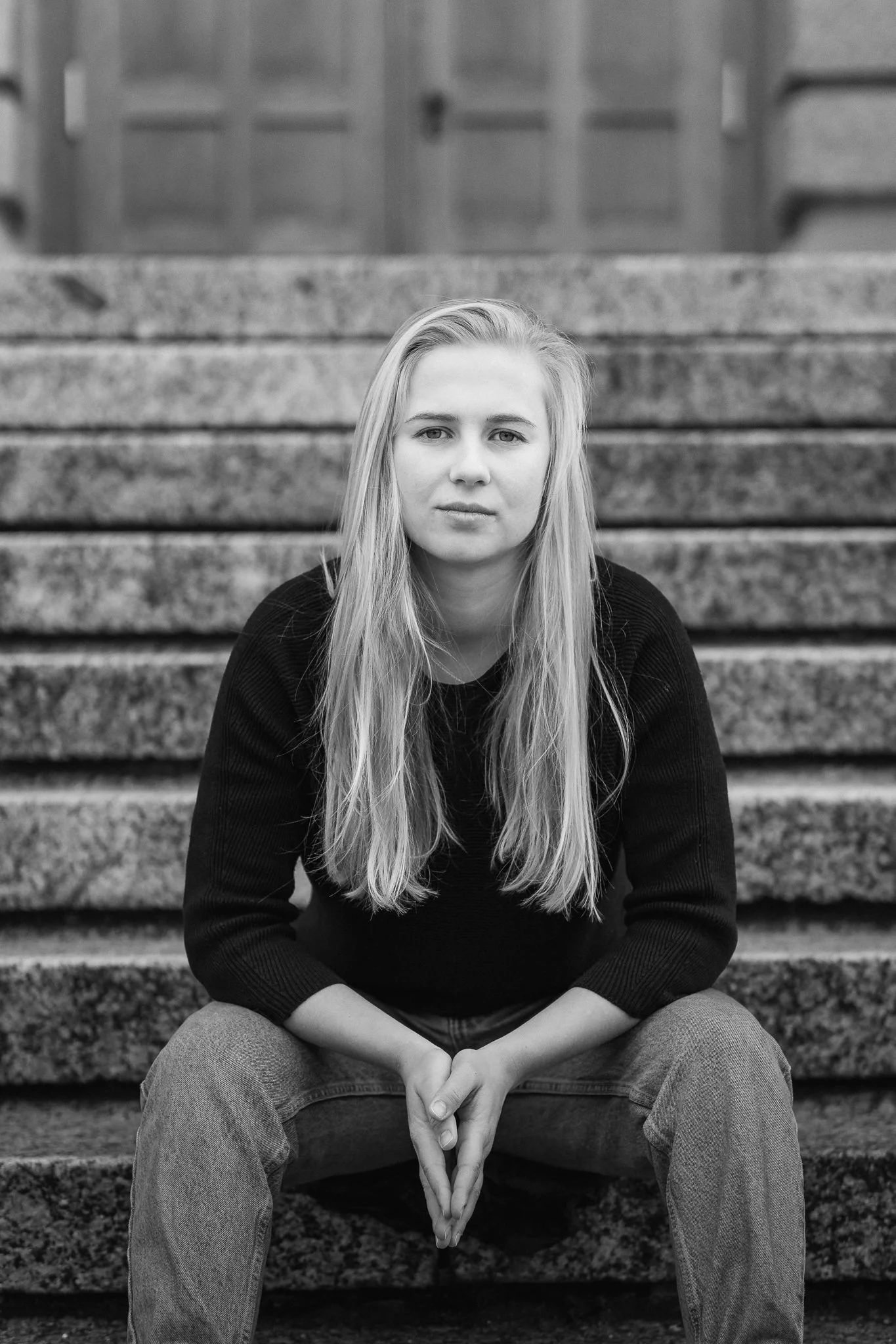 Junge Frau mit langen Haaren sitzend auf Steinstufen, im Hintergrund eine Holztür, in Schwarz-Weiß aufgenommen.