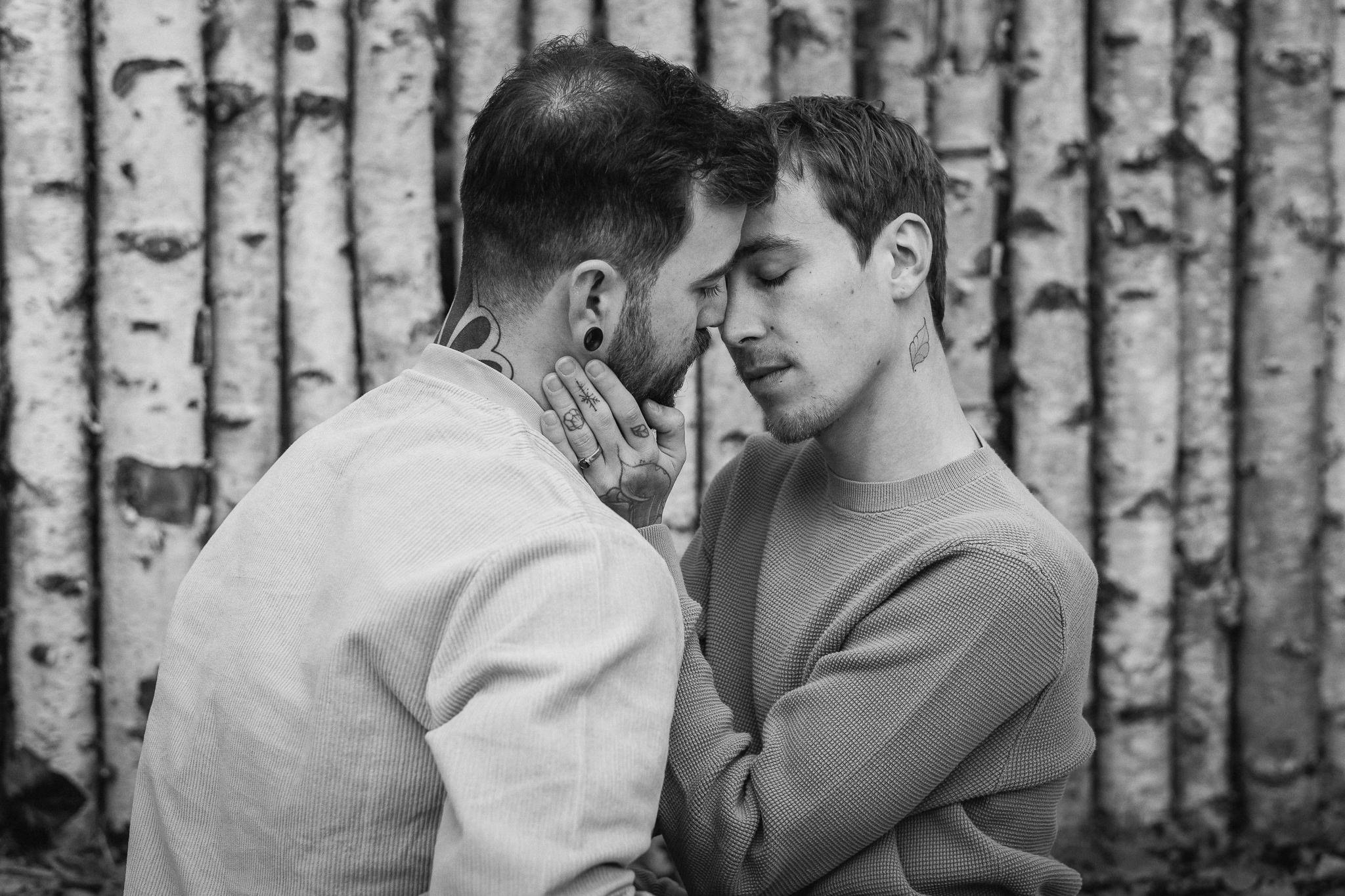 Zwei junge Männer stehen sich nah, berühren ihre Stirnen und halten sich an den Händen vor einer Baumrückwand, in Schwarz-Weiß aufgenommen.