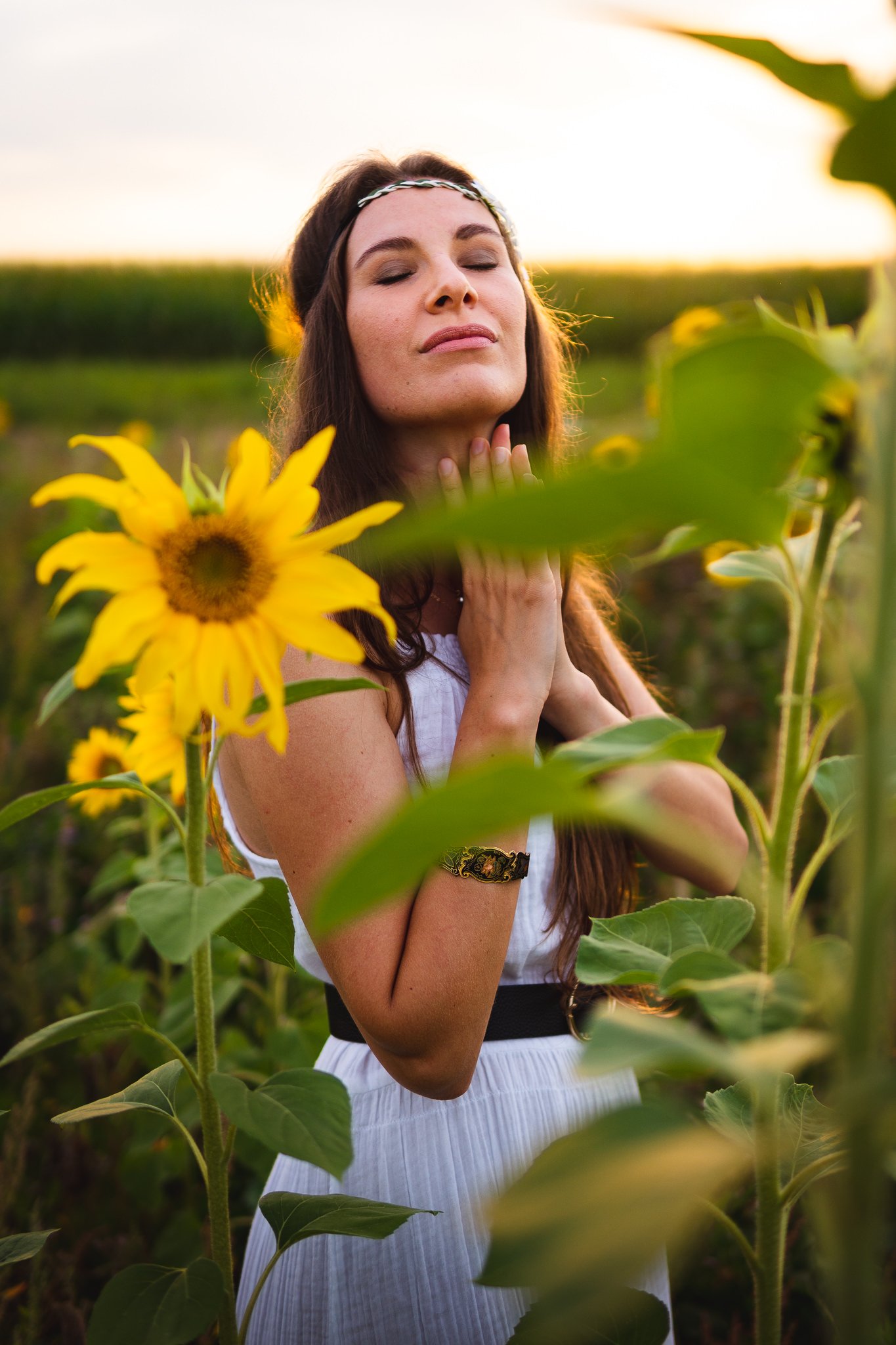 Eine junge Frau mit geschlossenen Augen steht zwischen Sonnenblumen im Sonnenuntergang. Sie trägt ein weißes Kleid und ein Armband.