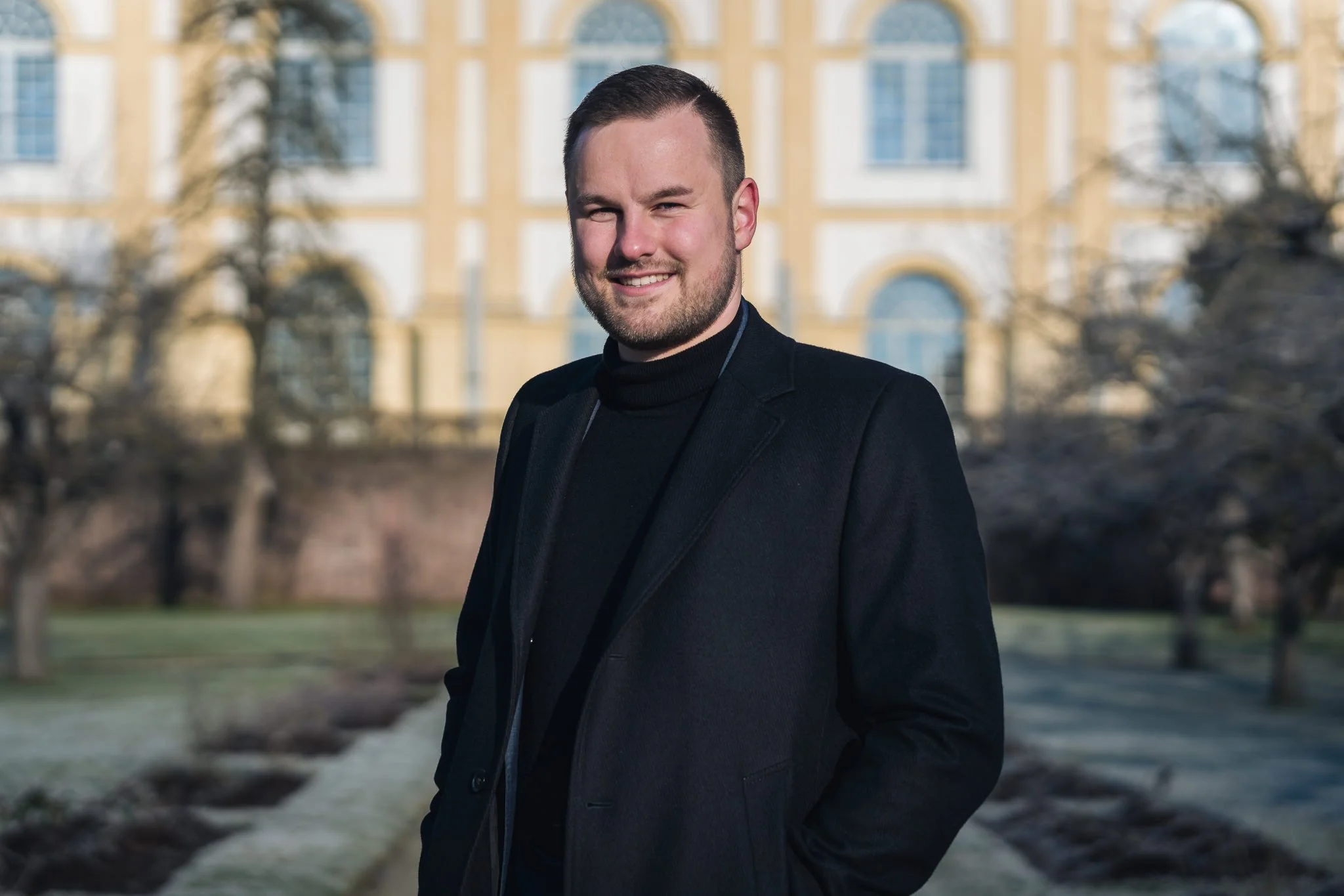 Ein junger Mann mit dunklem Haar und Bart, trägt eine schwarze Jacke und einen schwarzen Rollkragenpullover, steht im Freien vor einem historischen Gebäude mit großen Fenstern.