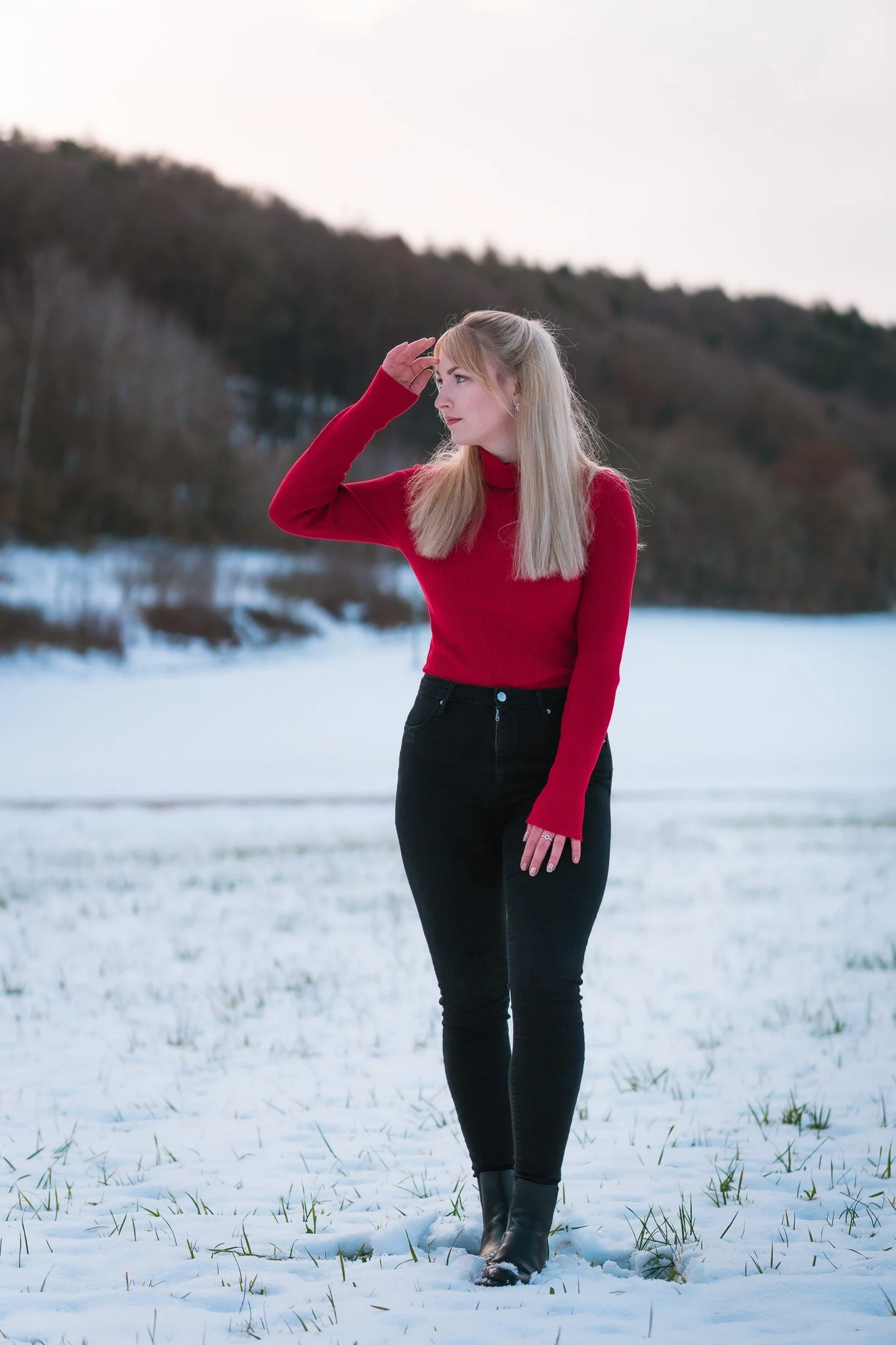 Junge Frau steht im Schnee in einer winterlichen Landschaft, trägt einen roten Pullover, schwarze Jeans und schwarze Stiefel, schaut nach rechts und berührt ihre Stirn mit der Hand.