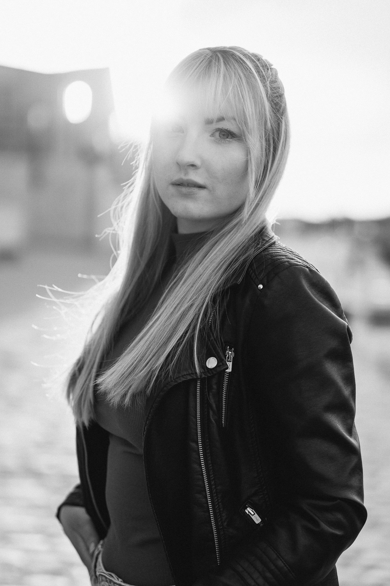 Schwarz-Weiß-Fotografie eines jungen Mädchens mit langem, glattem Haar, das eine Lederjacke trägt, im Freien bei Sonnenuntergang.