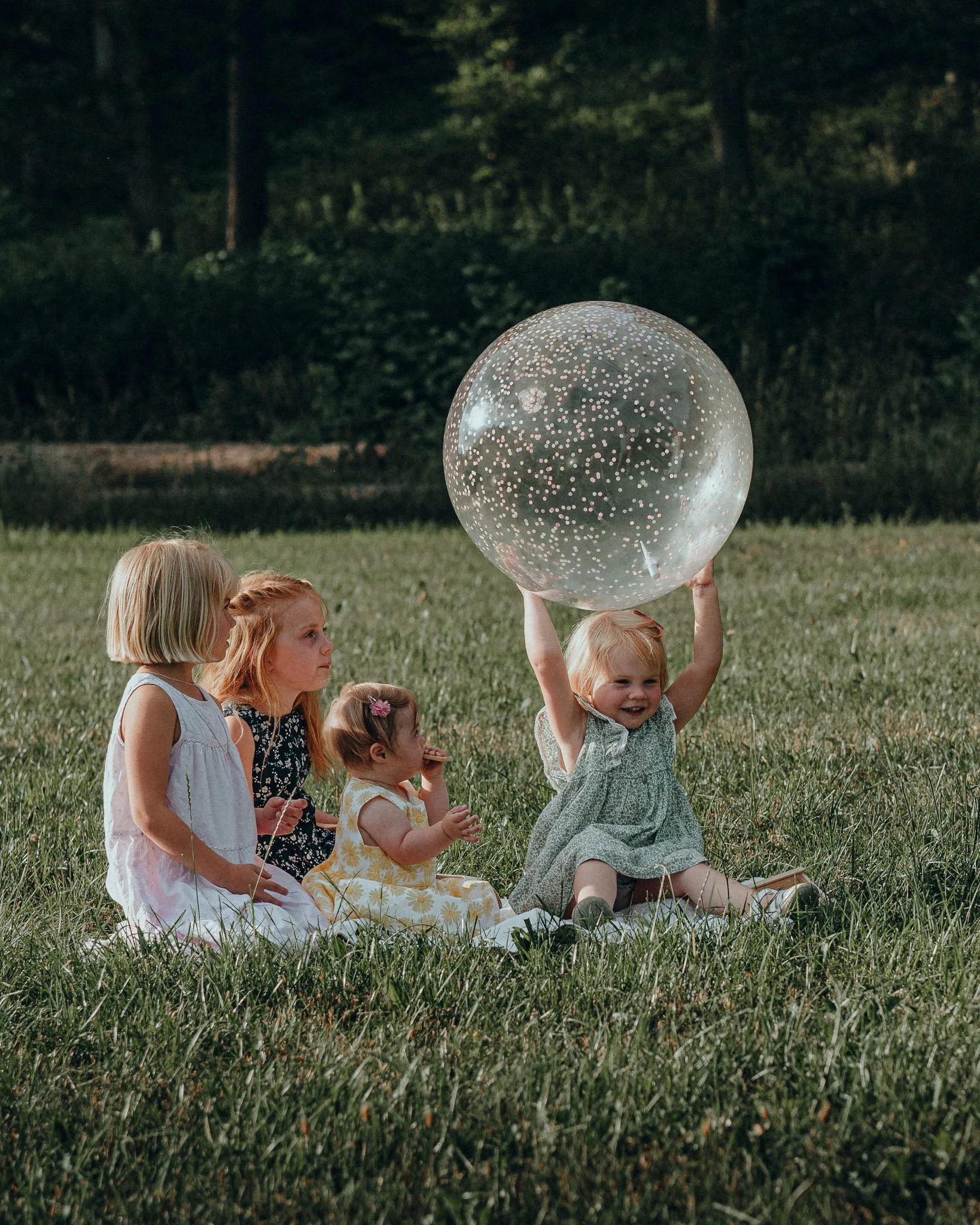 Vier Kinder sitzen auf einer Wiese, eine hält einen großen durchsichtigen Ball mit weißen Punkten über dem Kopf, während die anderen zuschauen.