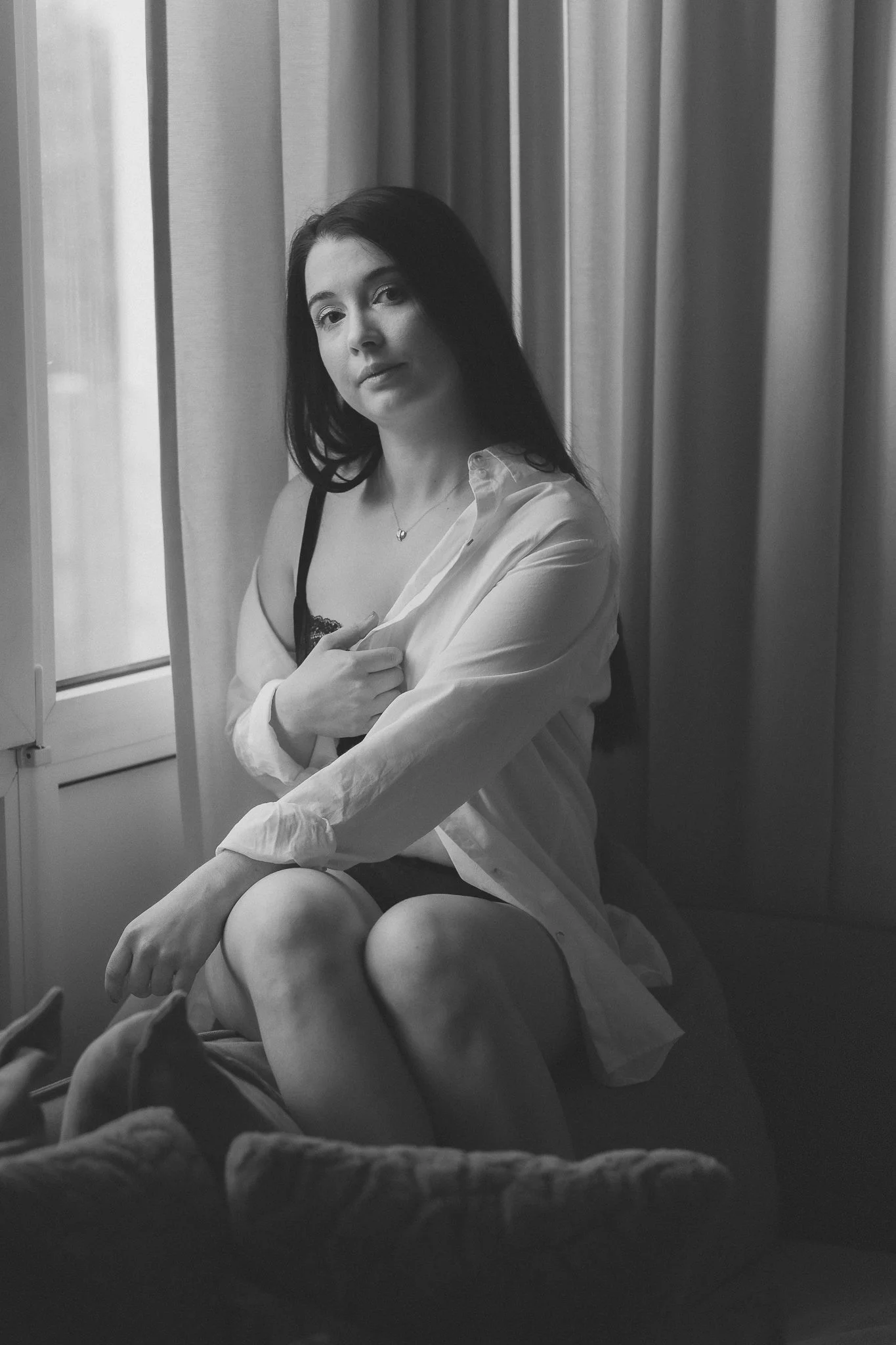 Schwarz-weiß-Foto eines sitzenden Mädchens vor einem Fenster mit Gardinen, trägt ein offenes Hemd und ein Unterhemd, schaut direkt in die Kamera.