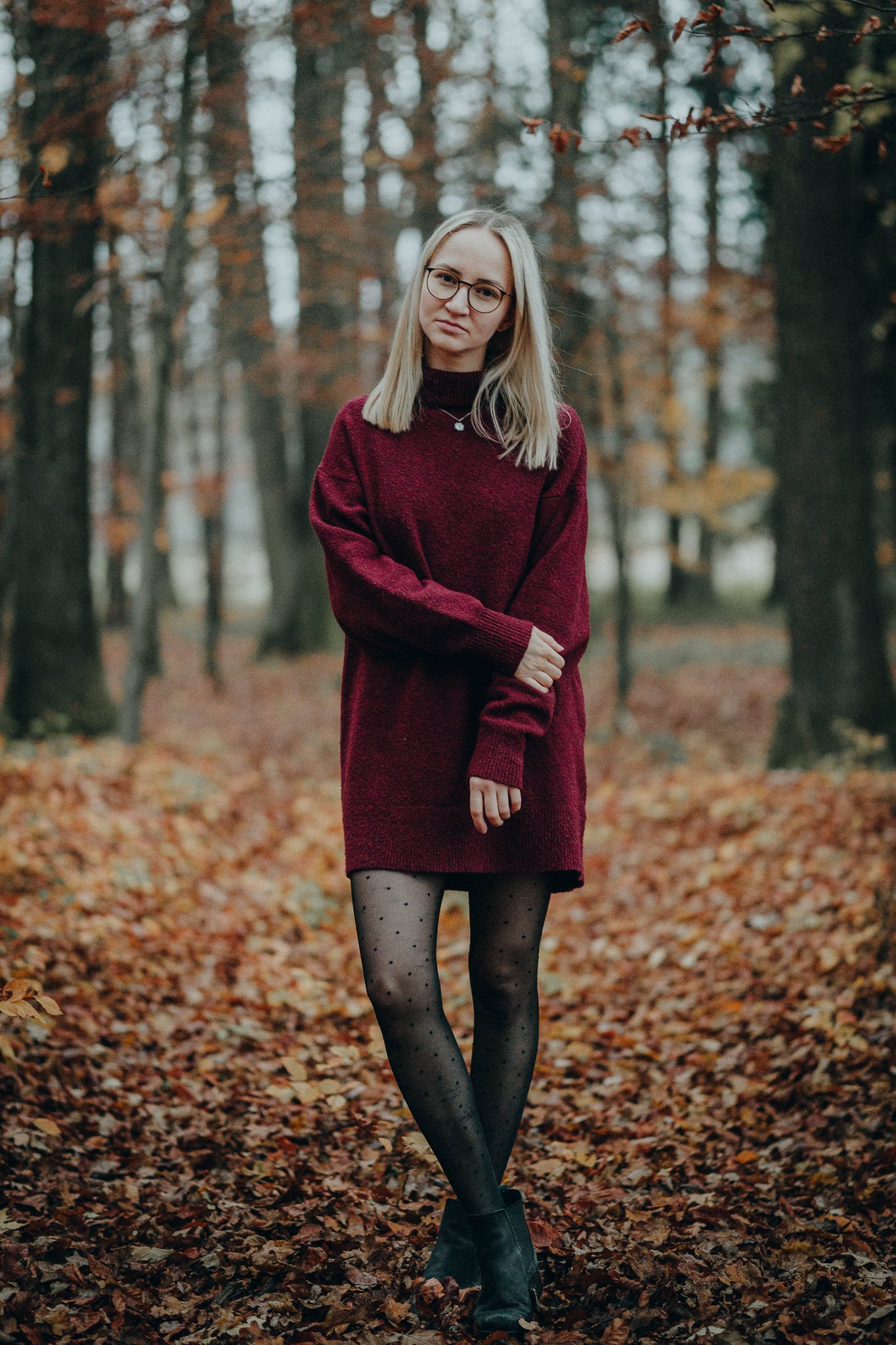 Junge Frau im Wald im Herbst, trägt eine rote Pulloverkleidung, schwarze Strumpfhosen mit Punkten, schwarze Schuhe, hat blondes Haar und trägt Brille.