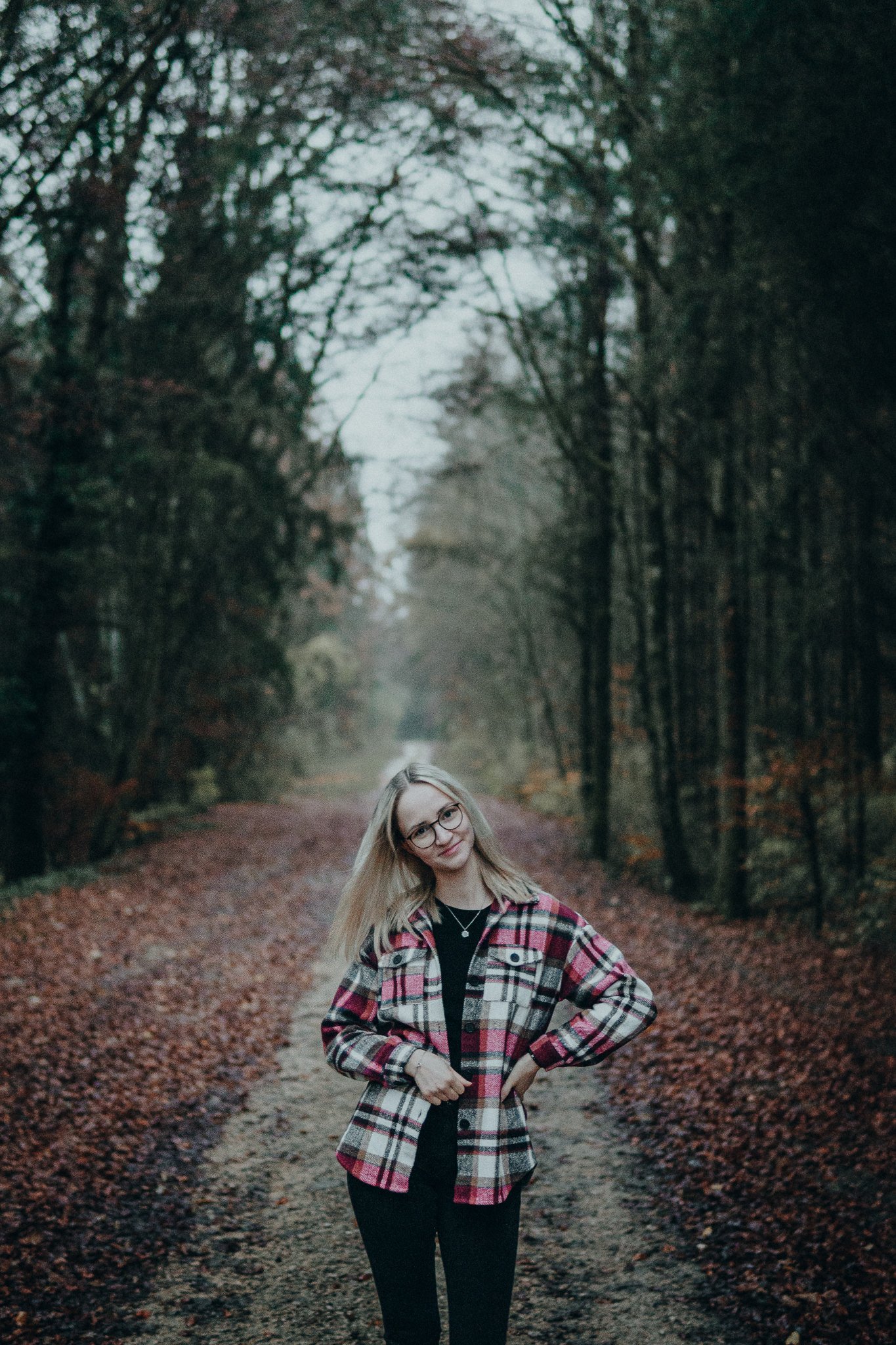 Eine junge Frau mit Brille und langem blondem Haar steht auf einem Weg in einem Wald im Herbst. Sie trägt eine bunte kariert Jacke und lächelt. Die Bäume sind ohne Blätter, der Himmel ist bewölkt.