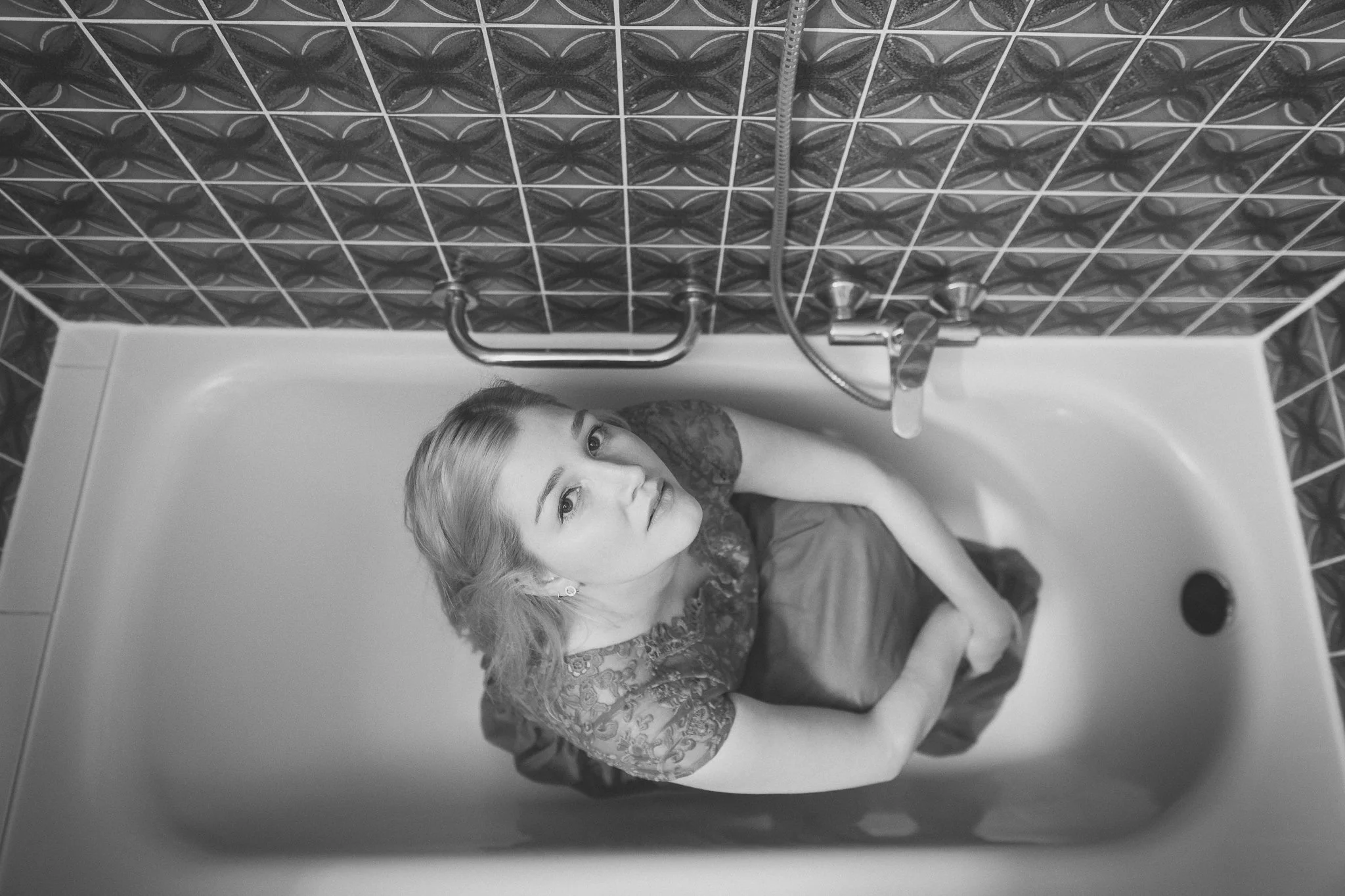 Schwarz-weiß-Foto einer jungen Frau mit blonden Haaren, die in einer Badewanne sitzt und nach oben schaut, umgeben von Wandfliesen mit geometrischem Muster.