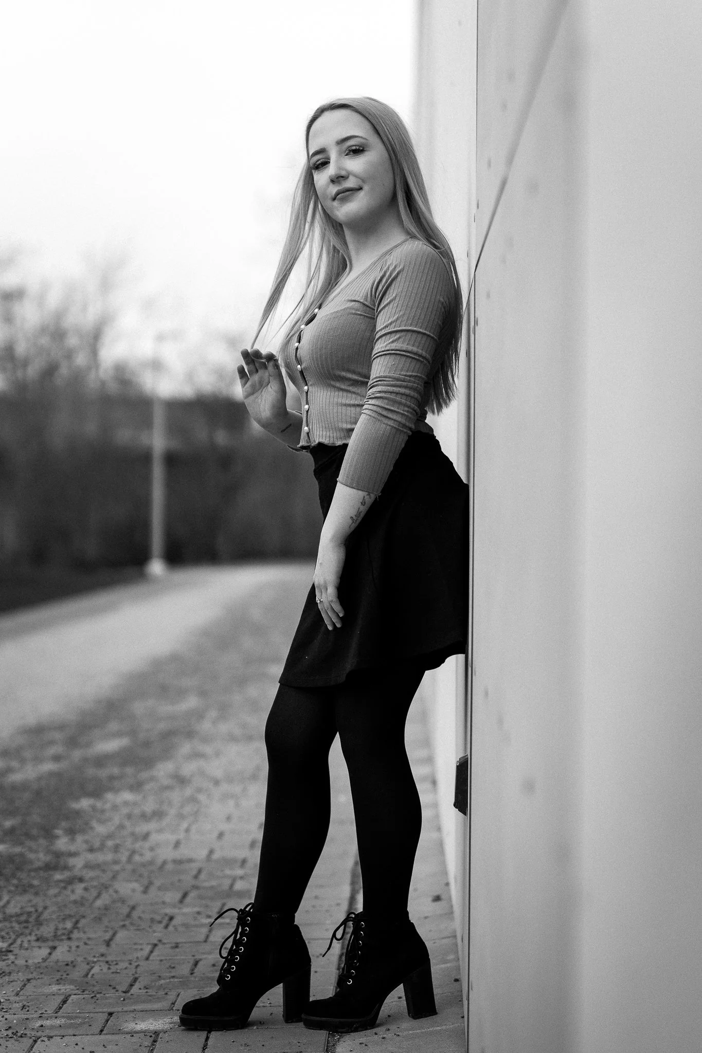 Schwarz-weisses Foto einer jungen Frau, die an einer Wand lehnt, mit langen Haaren, einem gerippten Top, Rock und hohen Schuhen, auf einer gepflasterten Straße, im Hintergrund Bäume und eine Laterne.