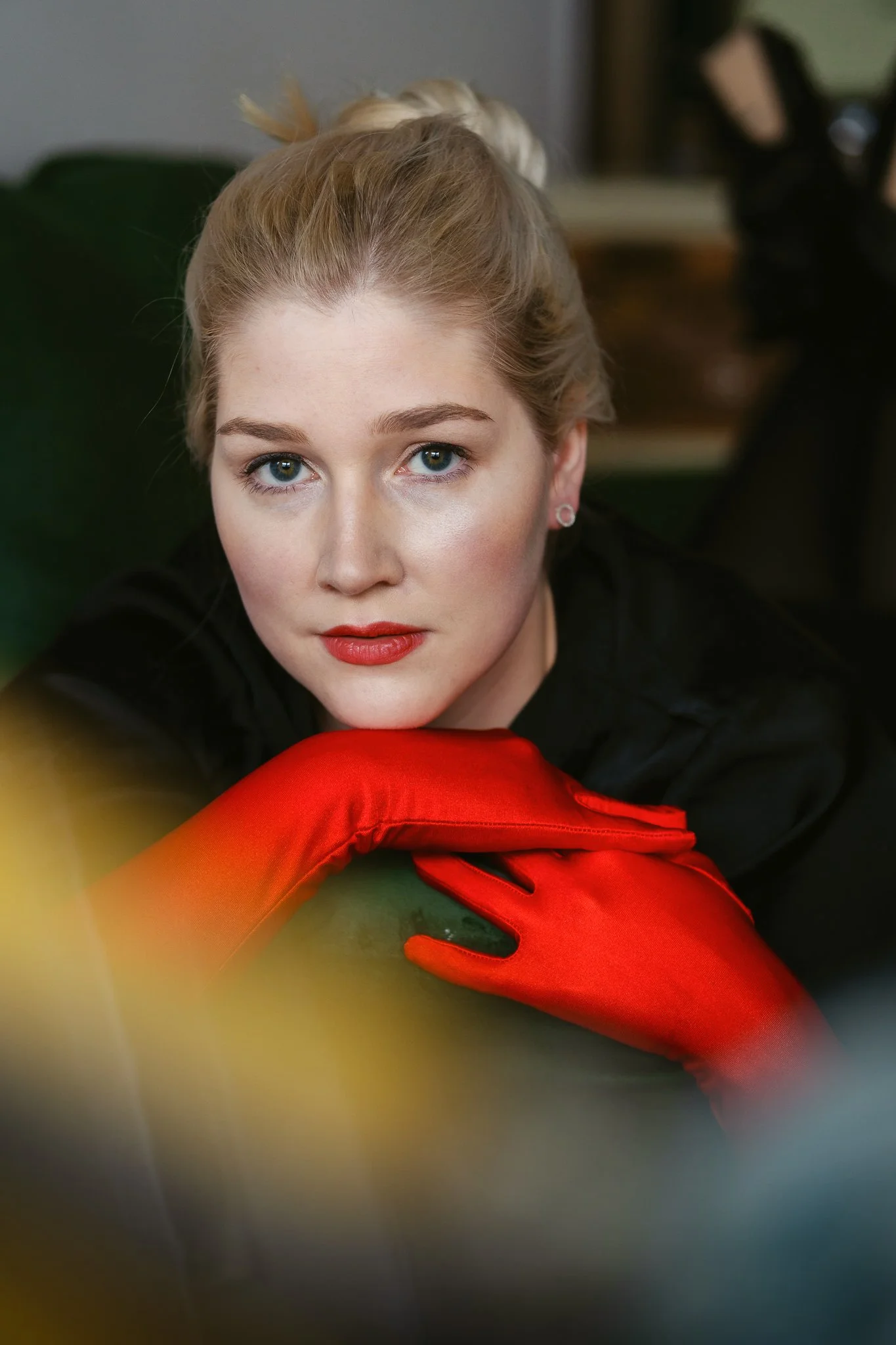 Nahaufnahme einer jungen Frau mit blonden Haaren, rotem Lippenstift, in schwarzer Kleidung und roten Handschuhen, die auf einem grünen Sofa sitzt.