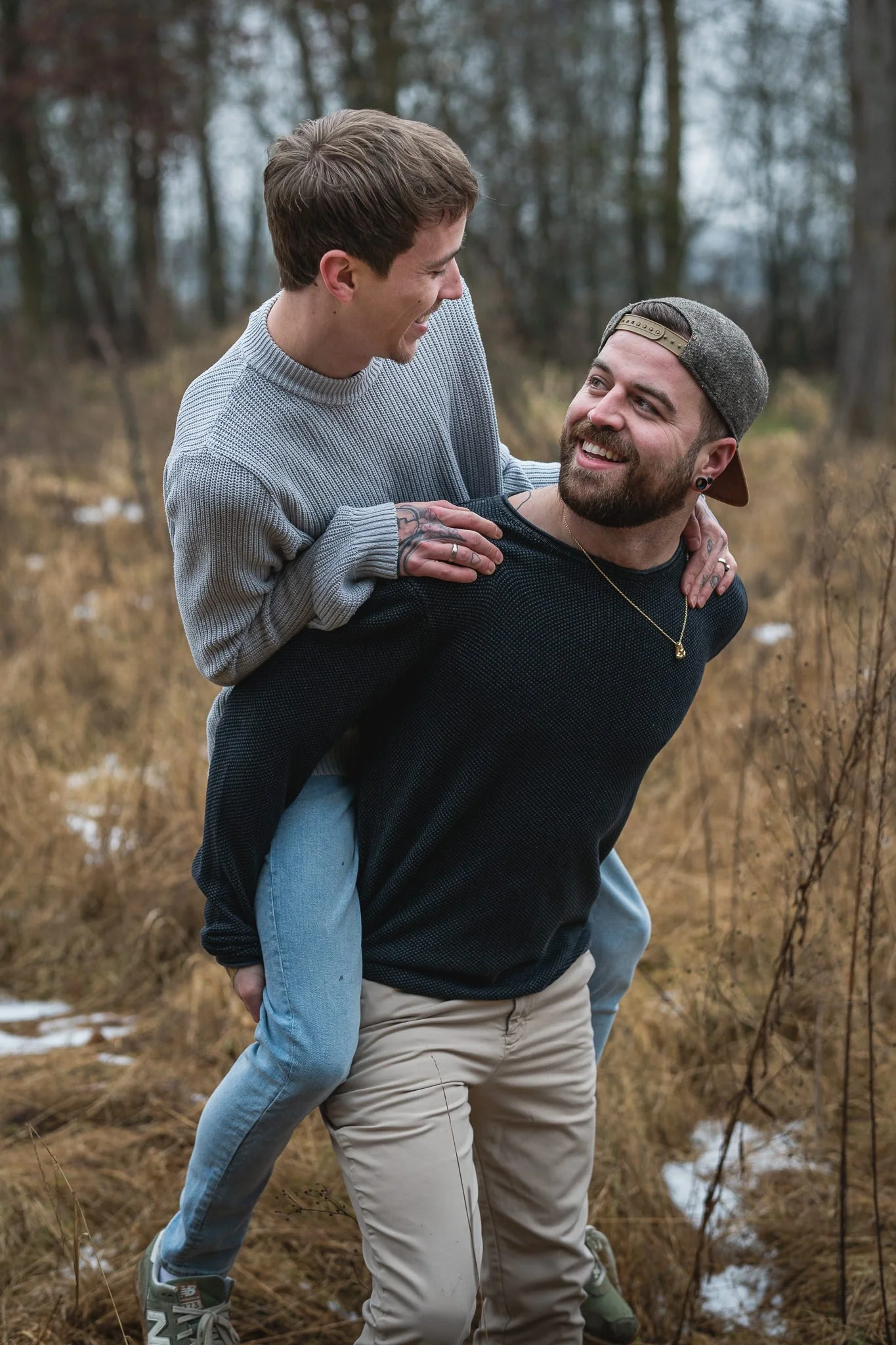 Zwei Männer genießen die Natur, der eine trägt den anderen auf dem Rücken durch ein trockenes, bewaldetes Gebiet mit wenig Schnee.