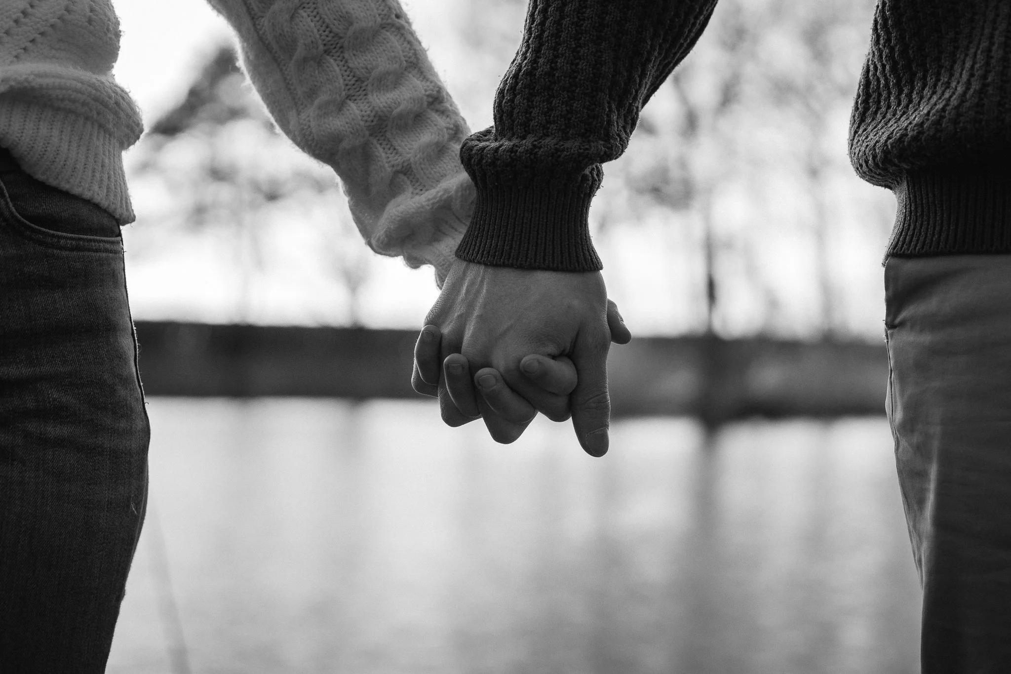 Zwei Personen halten sich an den Händen, draußen bei Wasser und Bäumen, schwarz-weiß