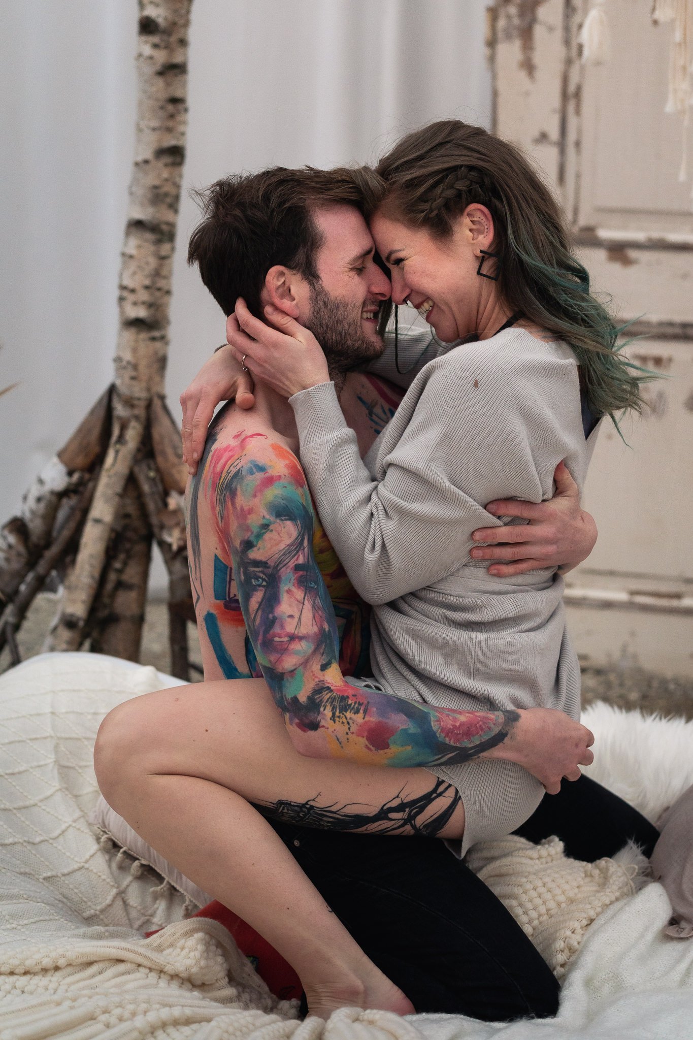 Paar umarmt sich lachend auf einem Bett, im Hintergrund Birkenstämme und eine alte Tür