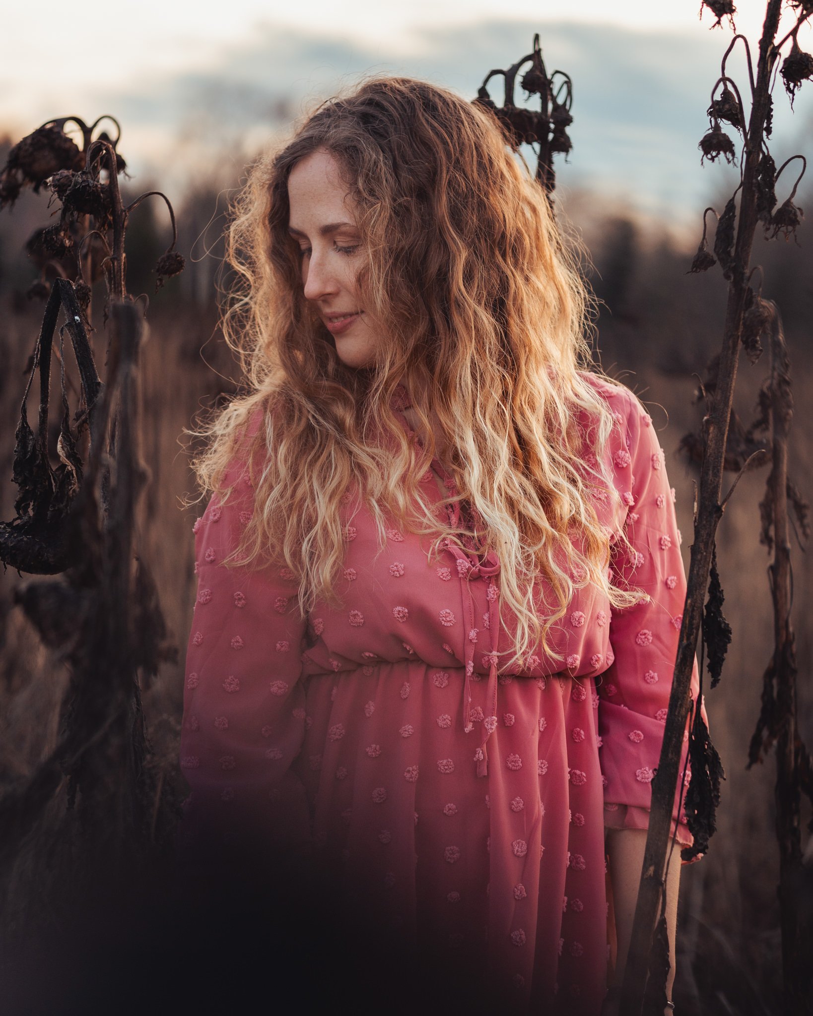 Eine Frau mit lockigem, hellbraunem Haar trägt ein rosa Kleid mit Stickerei und steht in einem Feld mit vertrockneten Pflanzen bei Sonnenuntergang.