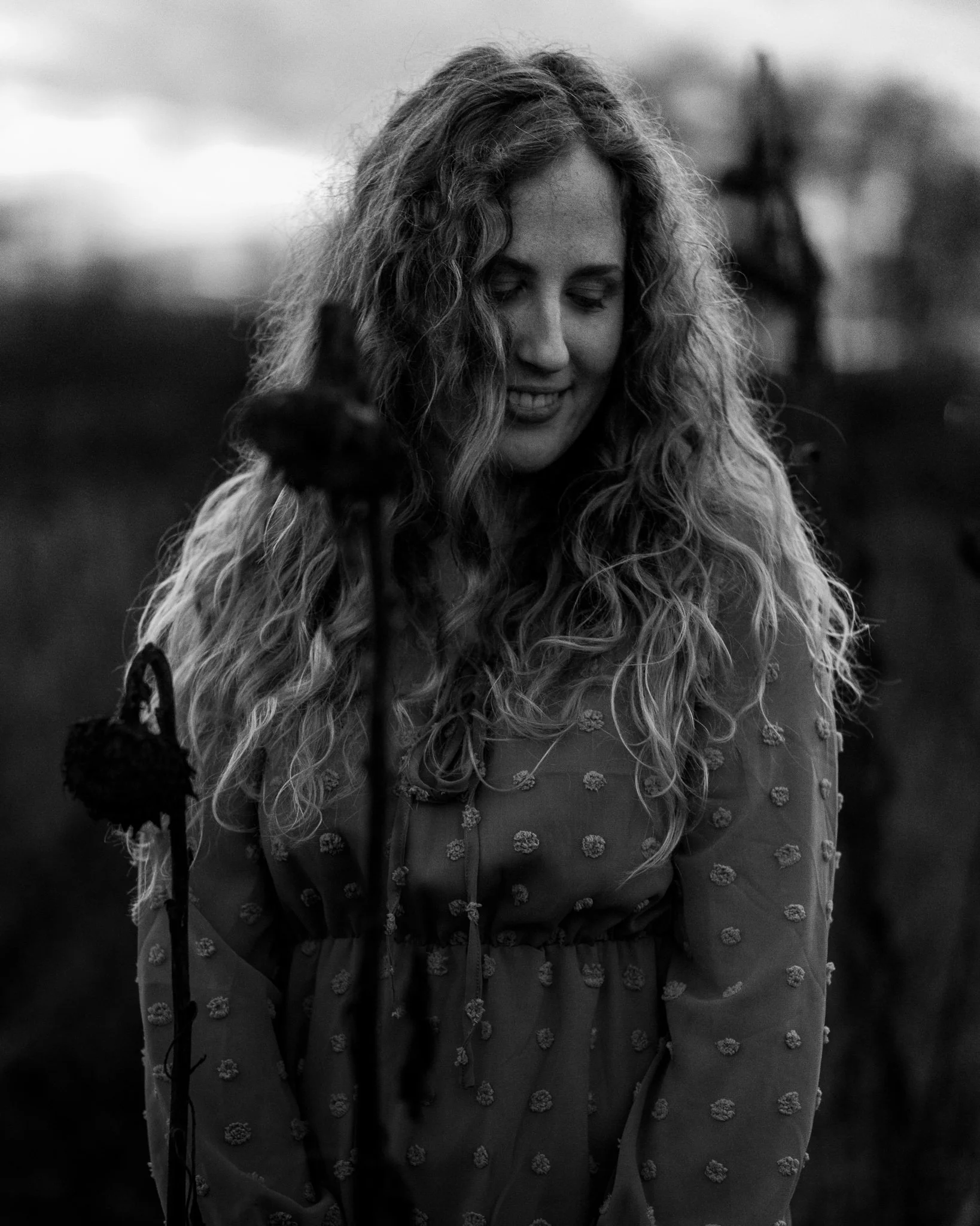 Schwarz-weiß-Foto eines lachenden Mädchens mit lockigem Haar, das ein Kleid mit Mustern trägt, im Freien bei Sonnenuntergang oder Dämmerung.