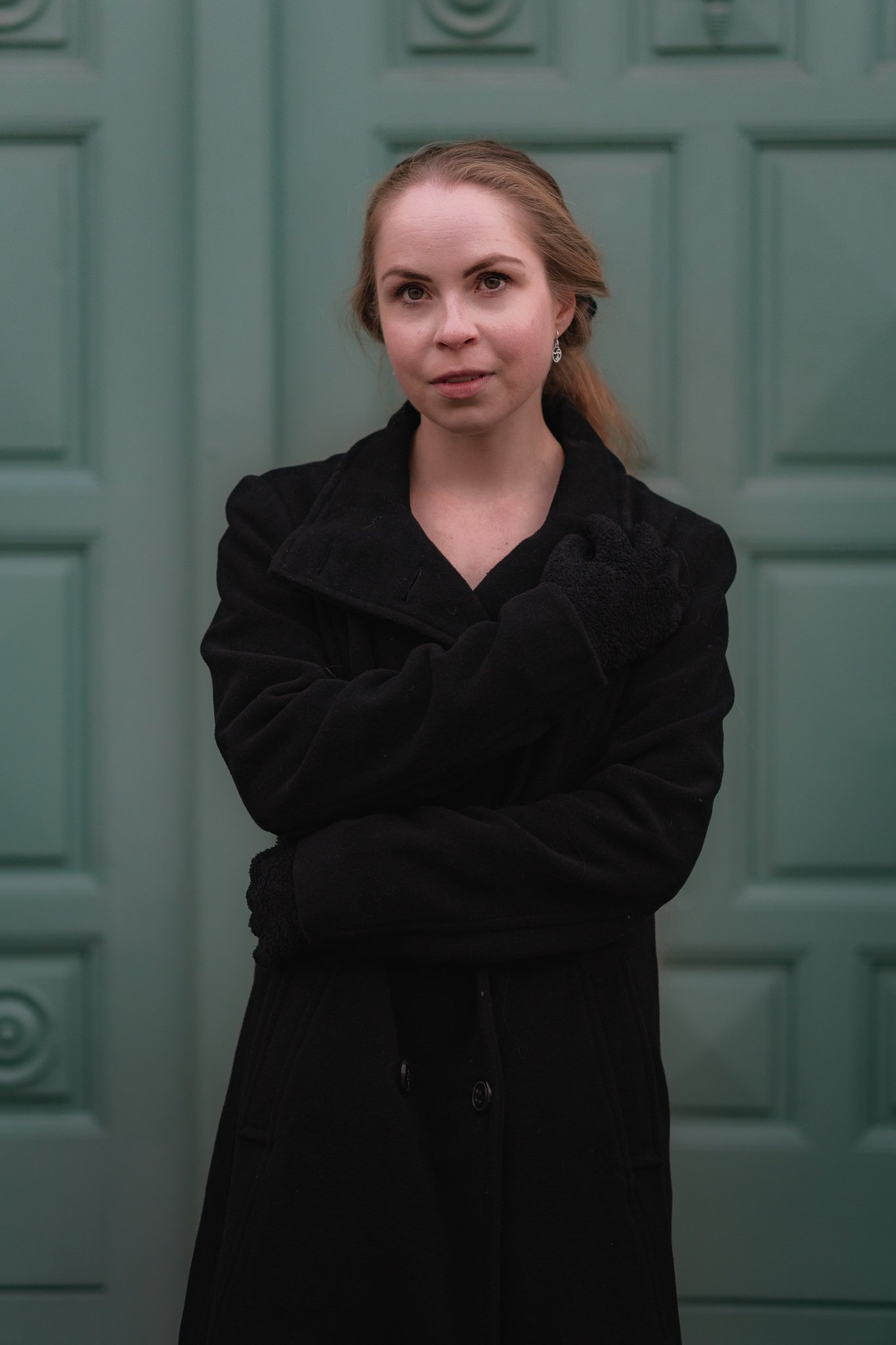 Junge Frau in schwarzem Mantel und Handschuhen vor grüner Tür.