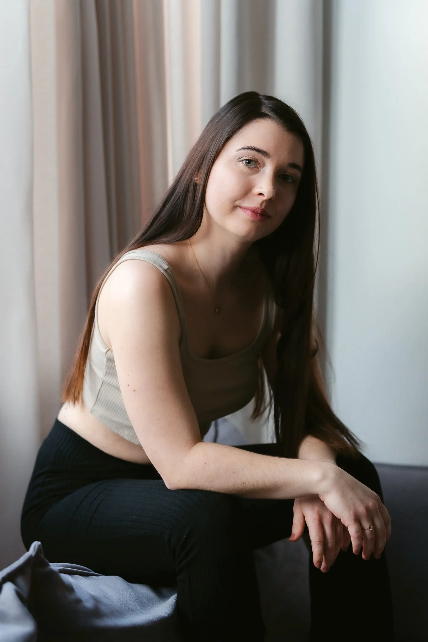 Junge Frau mit langen dunklen Haaren in einem beige Tanktop und schwarzen Hosen sitzt auf einem Sofa vor einem Fenster mit Vorhängen.