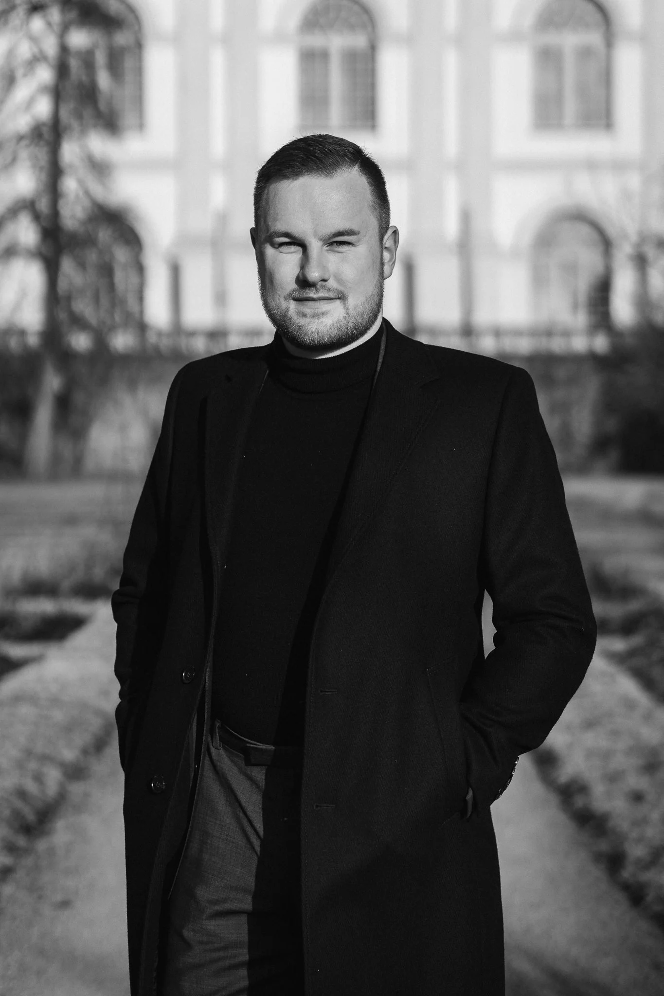 Ein junger Mann in dunkler Kleidung steht auf einem Weg vor einem großen Gebäude, im Hintergrund sind Bäume sichtbar.