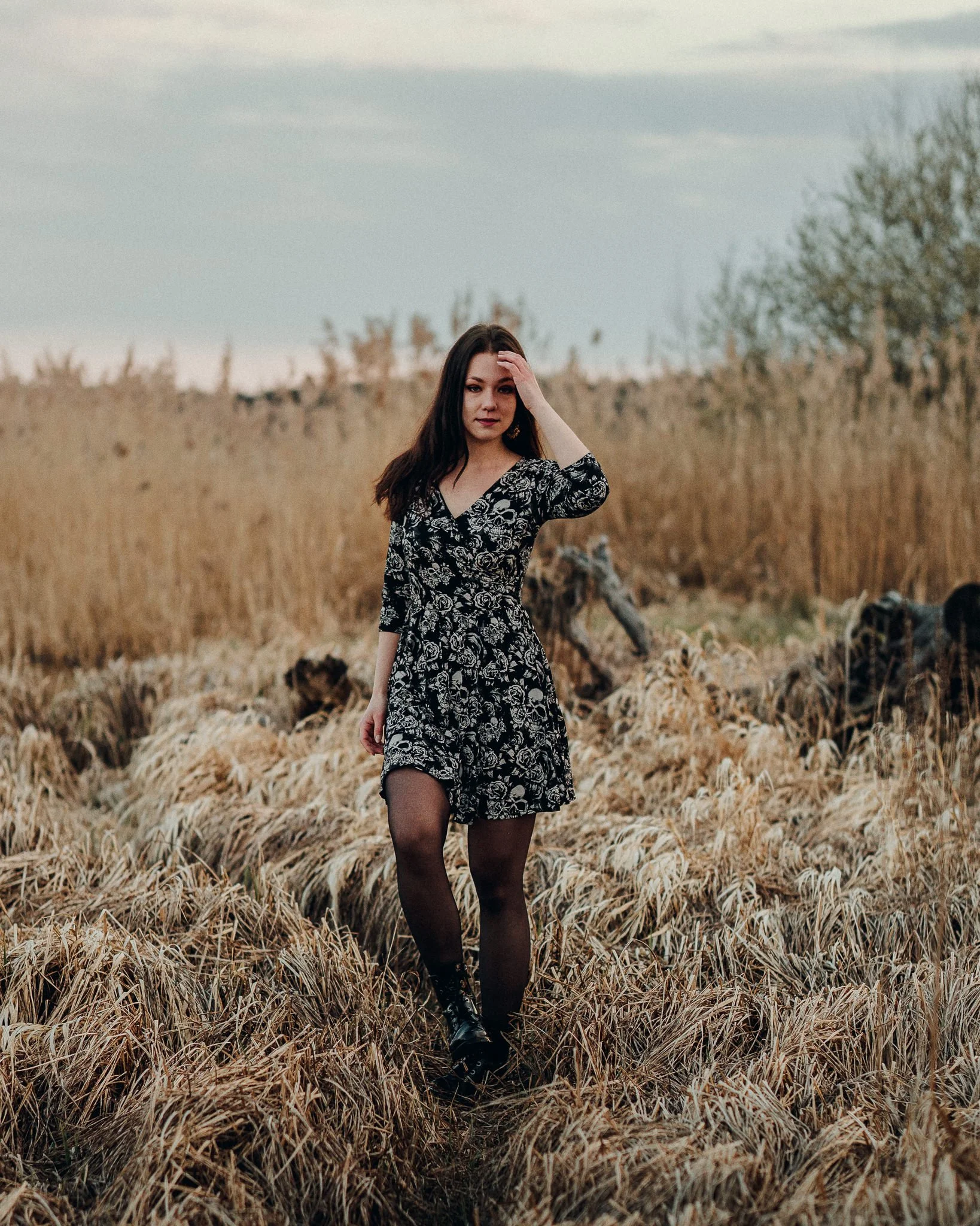 Eine junge Frau in einem schwarzen Kleid mit weißen Mustern steht in einem weiten Feld mit trockenen Gräsern, während sie eine Hand an den Kopf legt.