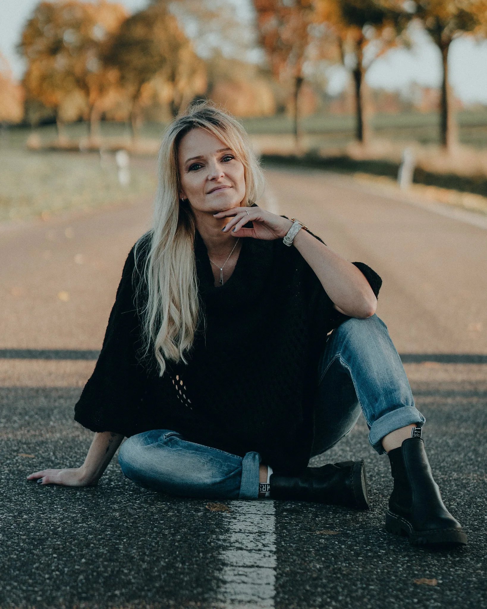 Frau sitzt auf einer Straße, umgeben von Bäumen mit Herbstlaub, trägt eine schwarze Strickjacke, Jeans und schwarze Stiefel, mit länglichem blondem Haar.