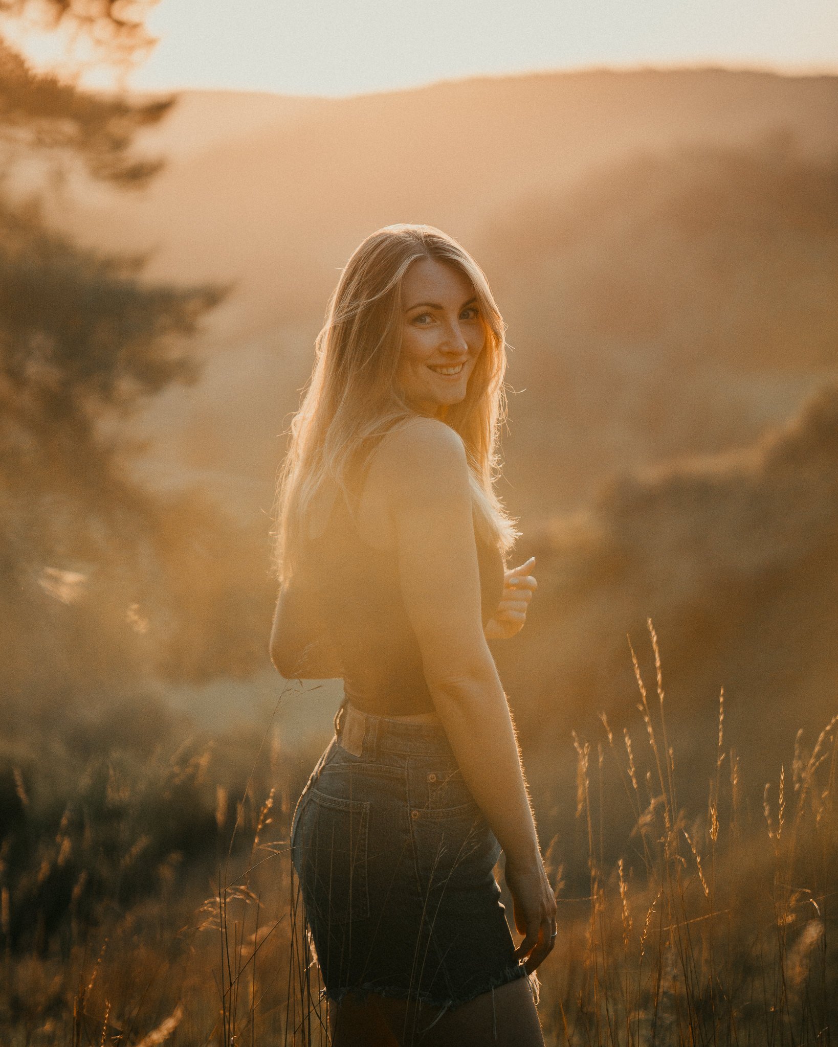 Eine lachende Frau in einem schwarzen Top und Jeansshorts steht in einem Weizenfeld bei Sonnenuntergang.