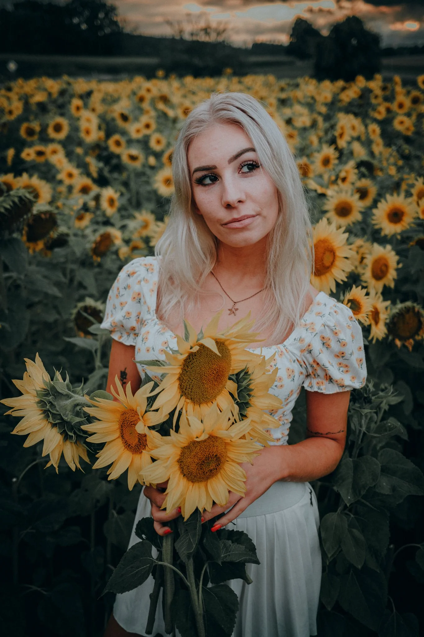 Eine blonde Frau steht inmitten eines Sonnenblumenfeldes bei Sonnenuntergang, hält einen großen Blumenstrauß mit Sonnenblumen und trägt ein weißes Kleid mit Blumenmuster.
