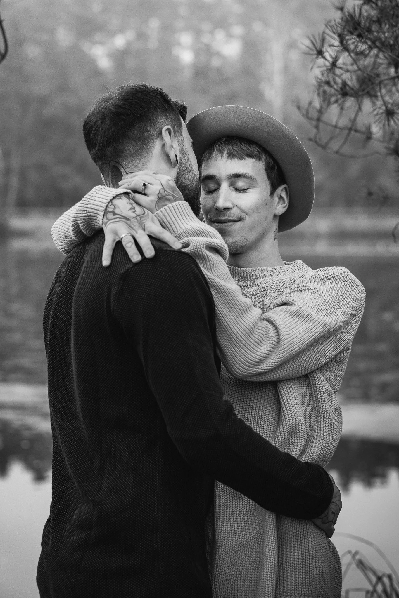 Zwei Männer umarmen sich liebevoll am See, einer trägt einen Hut, die andere hat Tattoos an den Händen, im Hintergrund Bäume und Wasser, schwarz-weiß.