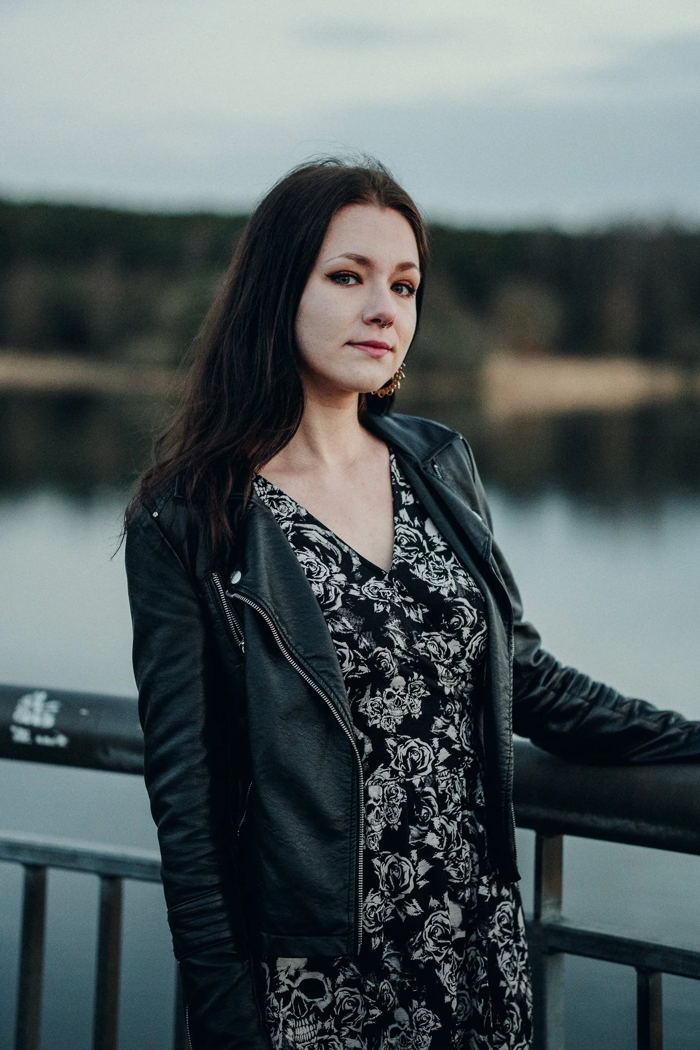 Junge Frau mit schwarzen Haaren, Lederjacke und Blumenkleid steht an einem Wasserufer und blickt in die Kamera.