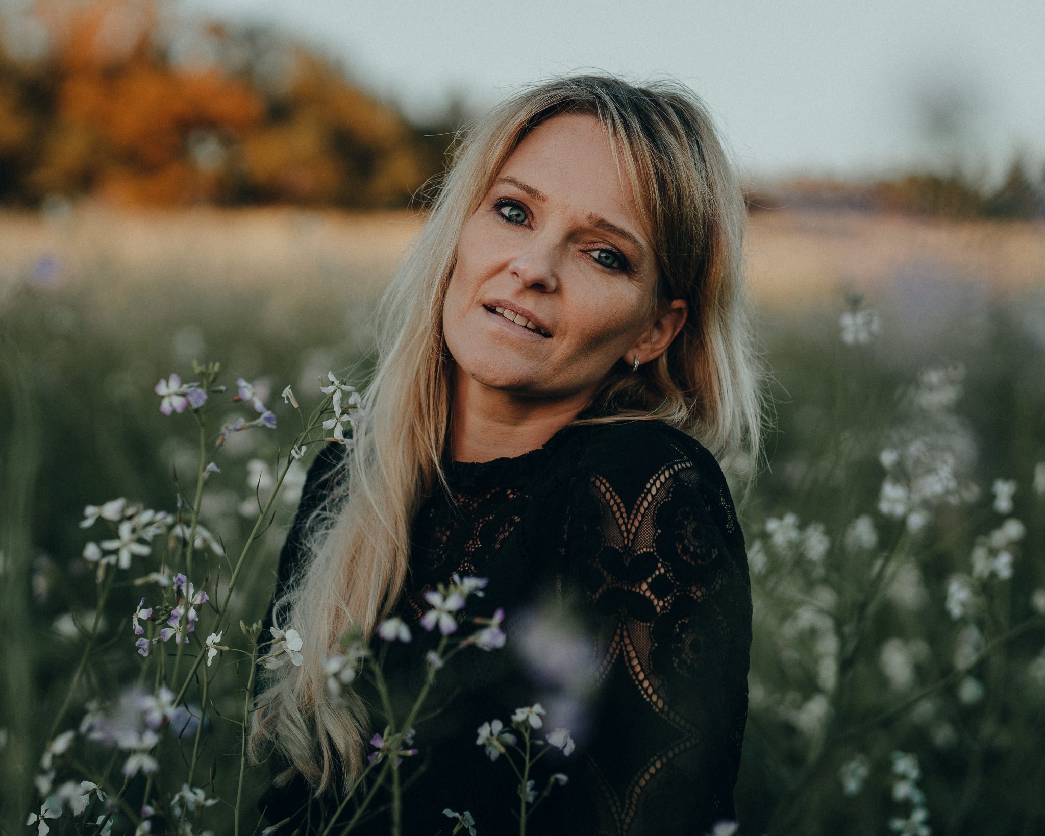 Ein lächelnder blonde Frau in einem schwarzen Spitzenkleid sitzt in einem Blumenfeld im Freien, im Hintergrund Bäume und ein Himmel bei Sonnenuntergang.