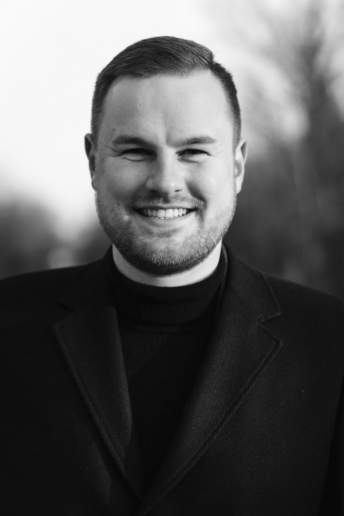 Schwarz-Weiß-Porträt eines lachenden Mannes mit kurz geschnittenem Haar und Bart, trägt einen schwarzen Anzug und Rollkragenpullover.
