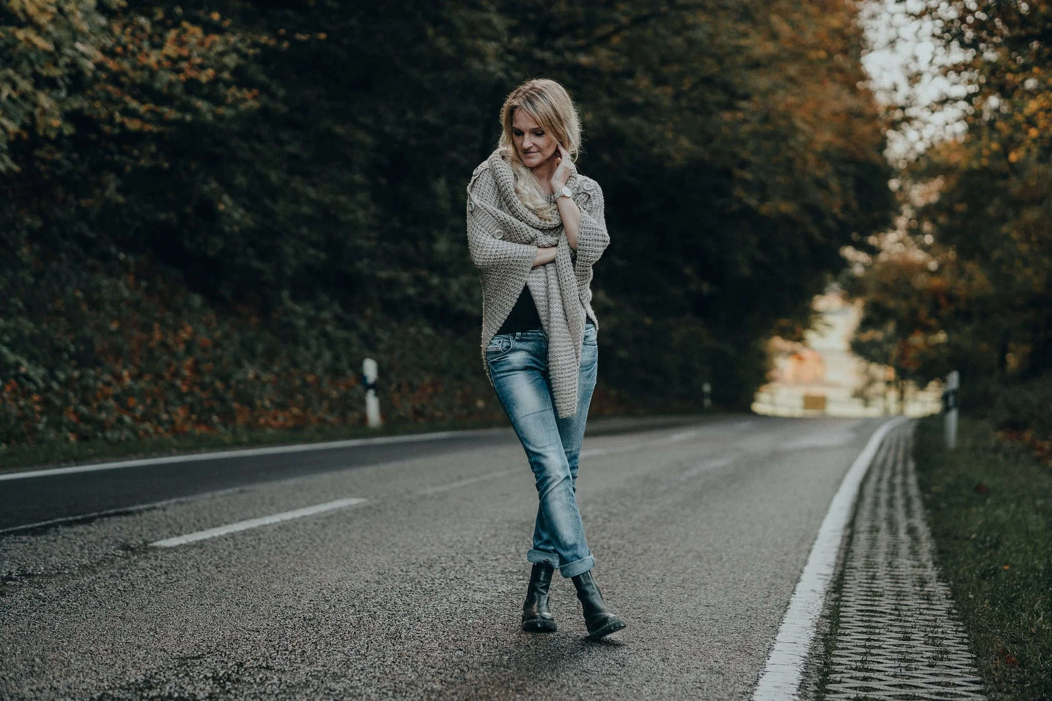 Frau läuft auf einer einsamen Straße umgeben von Bäumen im Herbst, trägt Jeans, Stiefel und einen grauen Mantel, wirkt nachdenklich.