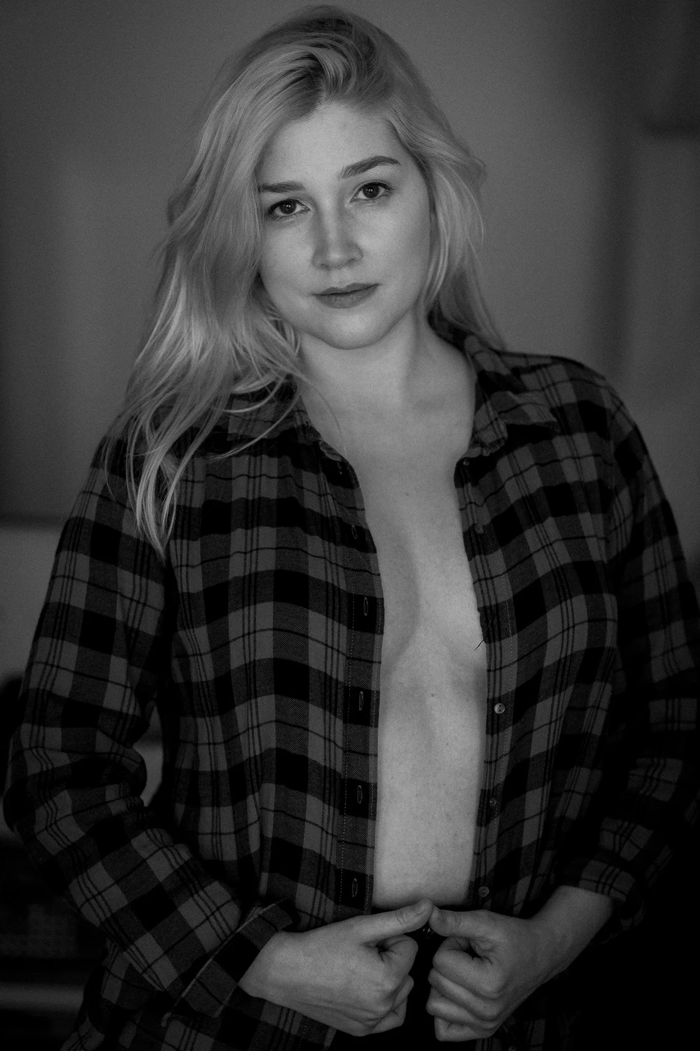 Schwarz-weiß-Foto einer jungen Frau mit lockigem, schulterlangem Haar, die eine offene, karierte Bluse trägt. Sie blickt in die Kamera, hält die unteren Knöpfe ihrer Bluse mit beiden Händen und zeigt damit teils ihren Oberkörper.
