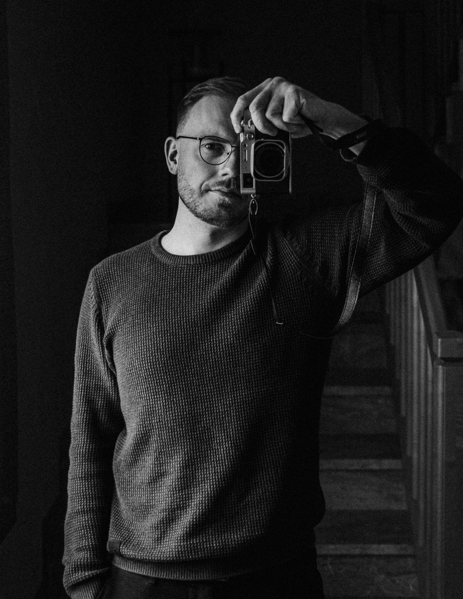 Mann macht Selbstporträt mit Kamera in Schwarz-Weiß, trägt Brille, dunkles Sweatshirt, steht vor dunklem Hintergrund.