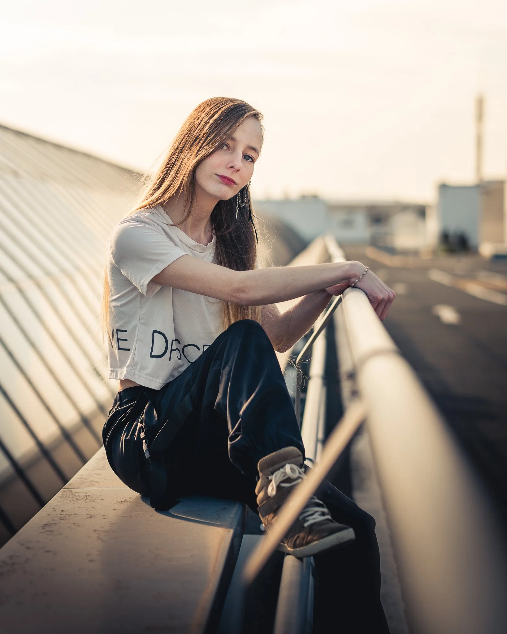 Junge Frau sitzt auf einem Flachdach, blickt nach unten, trägt ein weißes T-Shirt und schwarze Hosen, bei Sonnenuntergang.