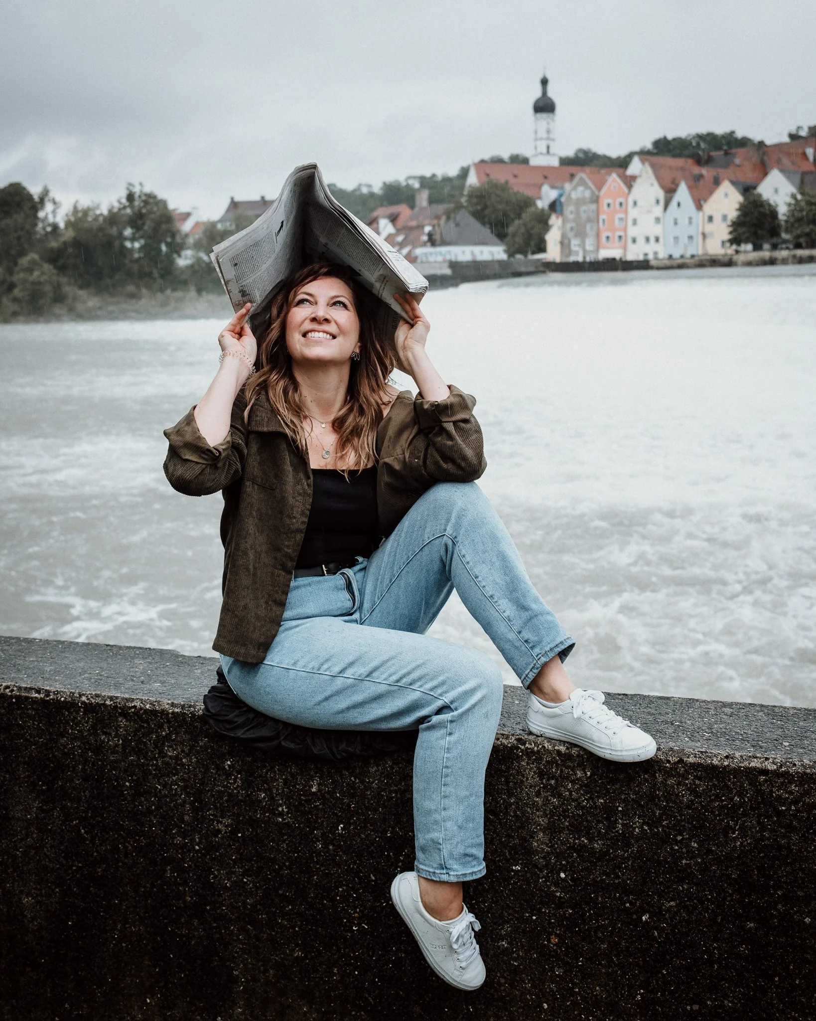 Eine junge Frau sitzt an einem Flussufer, hält eine Zeitung über den Kopf und blickt nach oben, mit einer malerischen Stadt im Hintergrund.