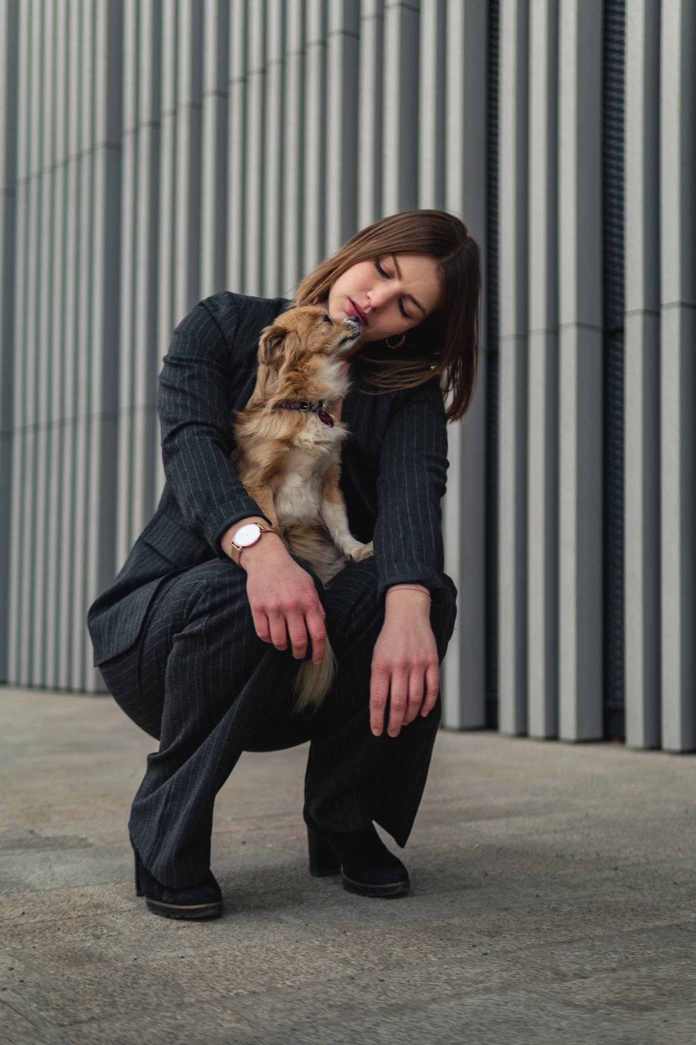 Eine Frau in einem schwarzen Anzug sitzt vor einer modernen Gebäudefassade, während sie mit einem kleinen braunen Hund interagiert, der sie liebevoll leckt.
