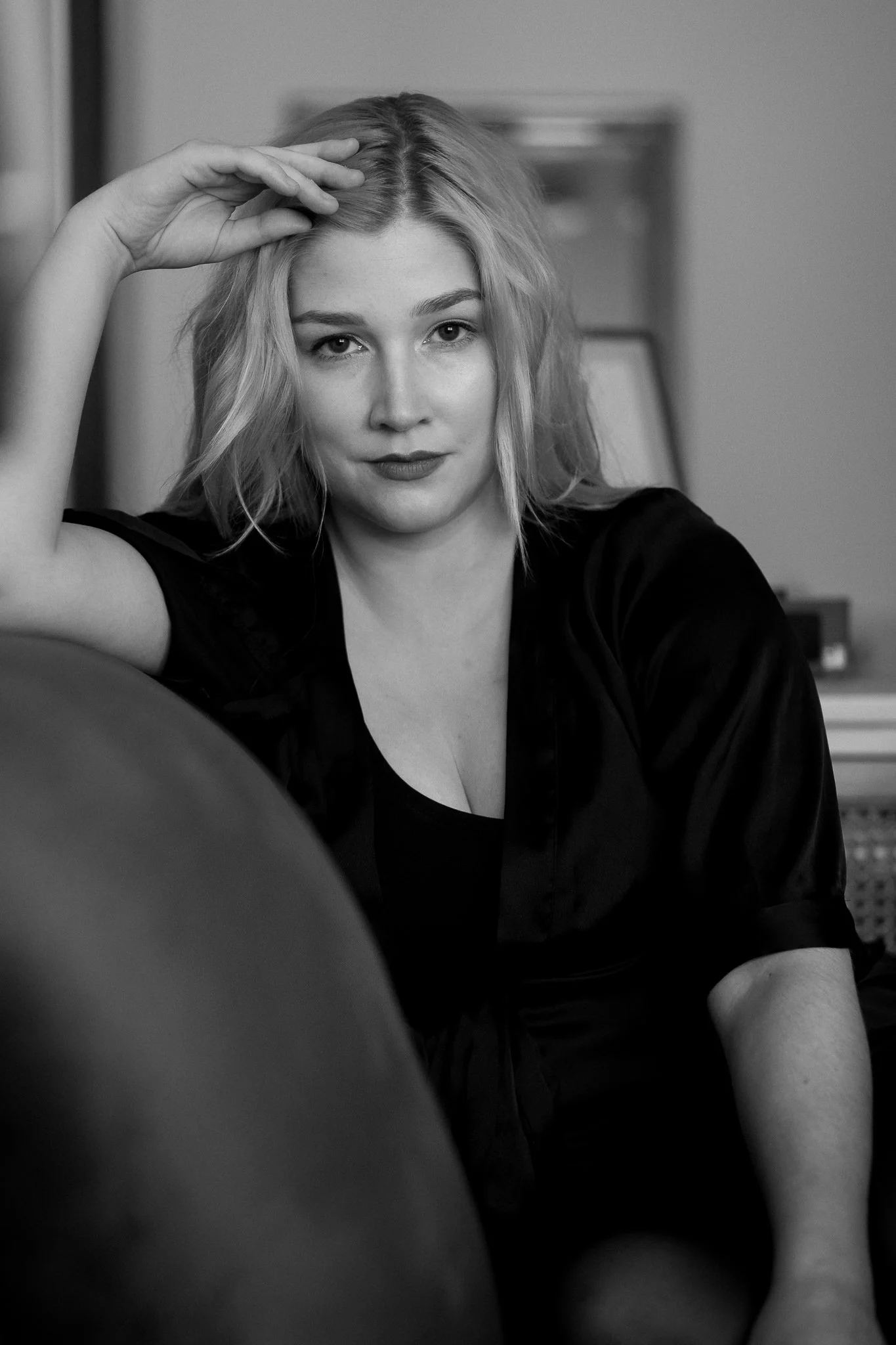 Schwarz-weiß-Foto eines jungen Mädchens mit blonden, lockigen Haaren, das auf einem Sofa sitzt. Sie trägt ein dunkles Oberteil und sieht direkt in die Kamera, lächelnd. Ihre Hand liegt auf ihrem Kopf, der Hintergrund ist unscharf mit Möbeln erkennbar