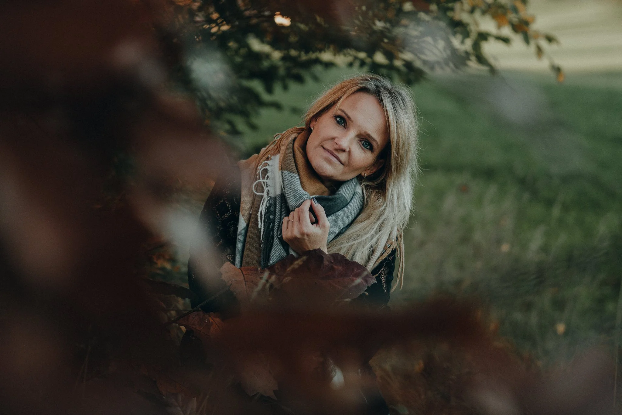 Eine blonde Frau in einem Waldgebiet, die einen either gestrickten Schal trägt und die Hand an die Brust legt.