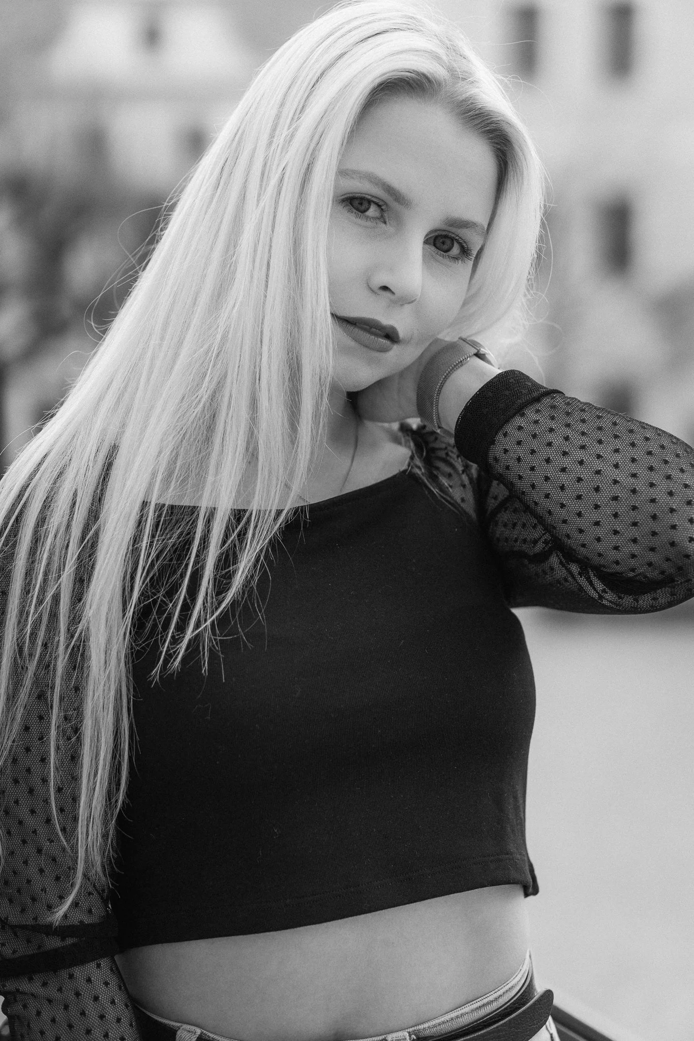 Schwarz-weiße Fotografie einer jungen Frau mit langen, hellen Haaren, die in die Kamera schaut, mit einem schwarzen Top mit transparenten, gepunkteten Ärmeln.