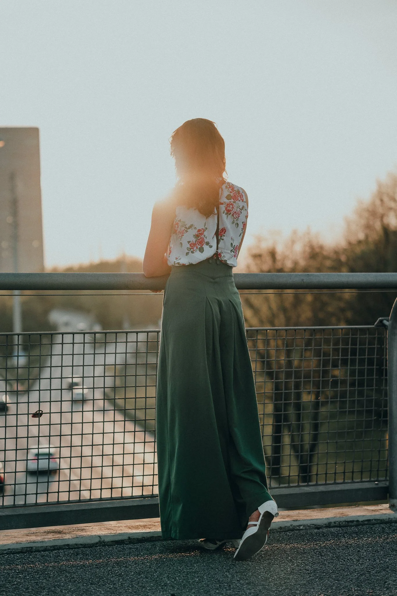Eine Frau, die an einem Geländer auf einer Brücke im Sonnenuntergang steht, trägt breite grüne Hosen und ein weißes Top mit Blumenmuster.