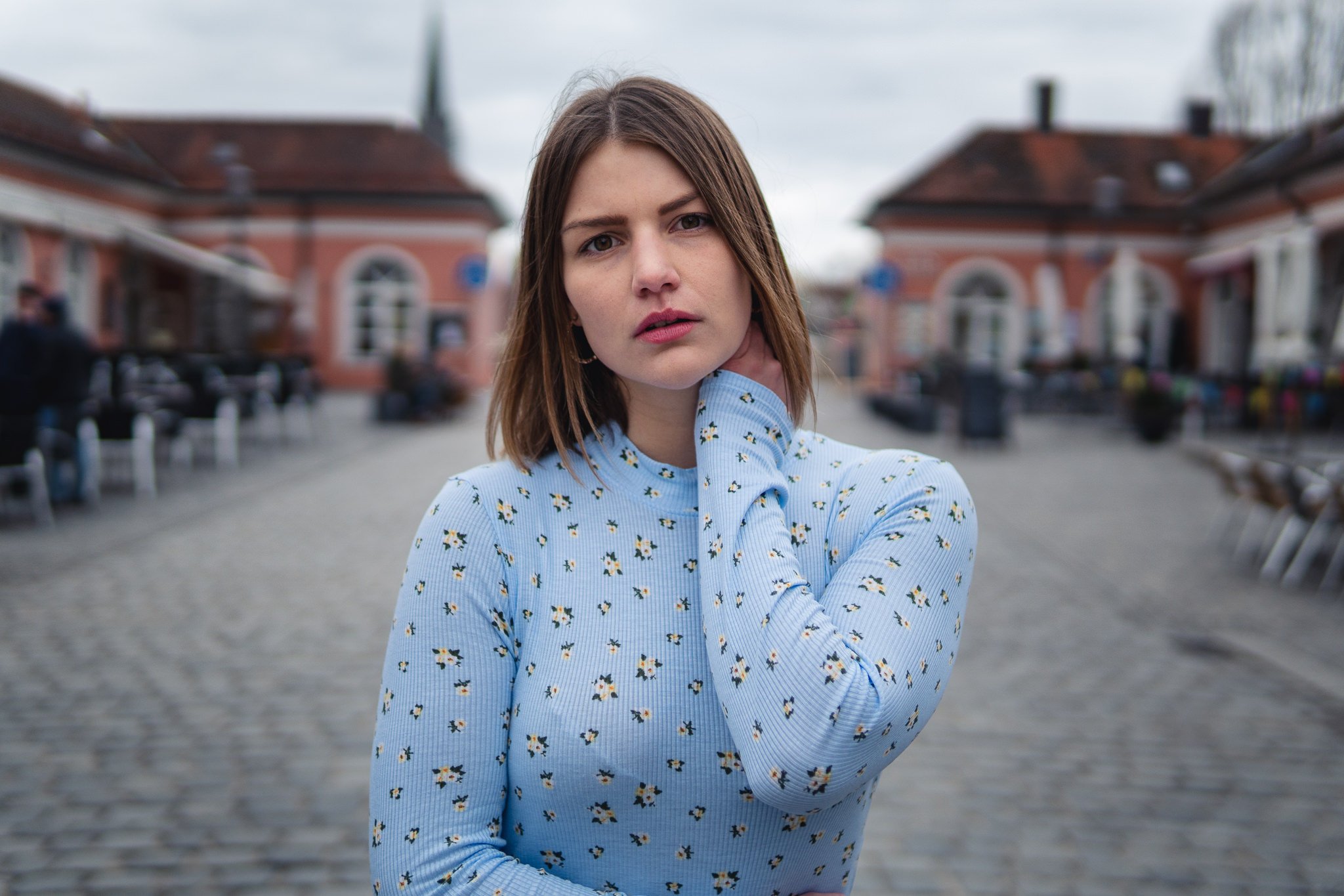 Junge Frau mit braunen Haaren in einem blauen Pullover mit Blumenmuster steht auf einer Kopfsteinpflasterstraße, im Hintergrund alte Gebäude.
