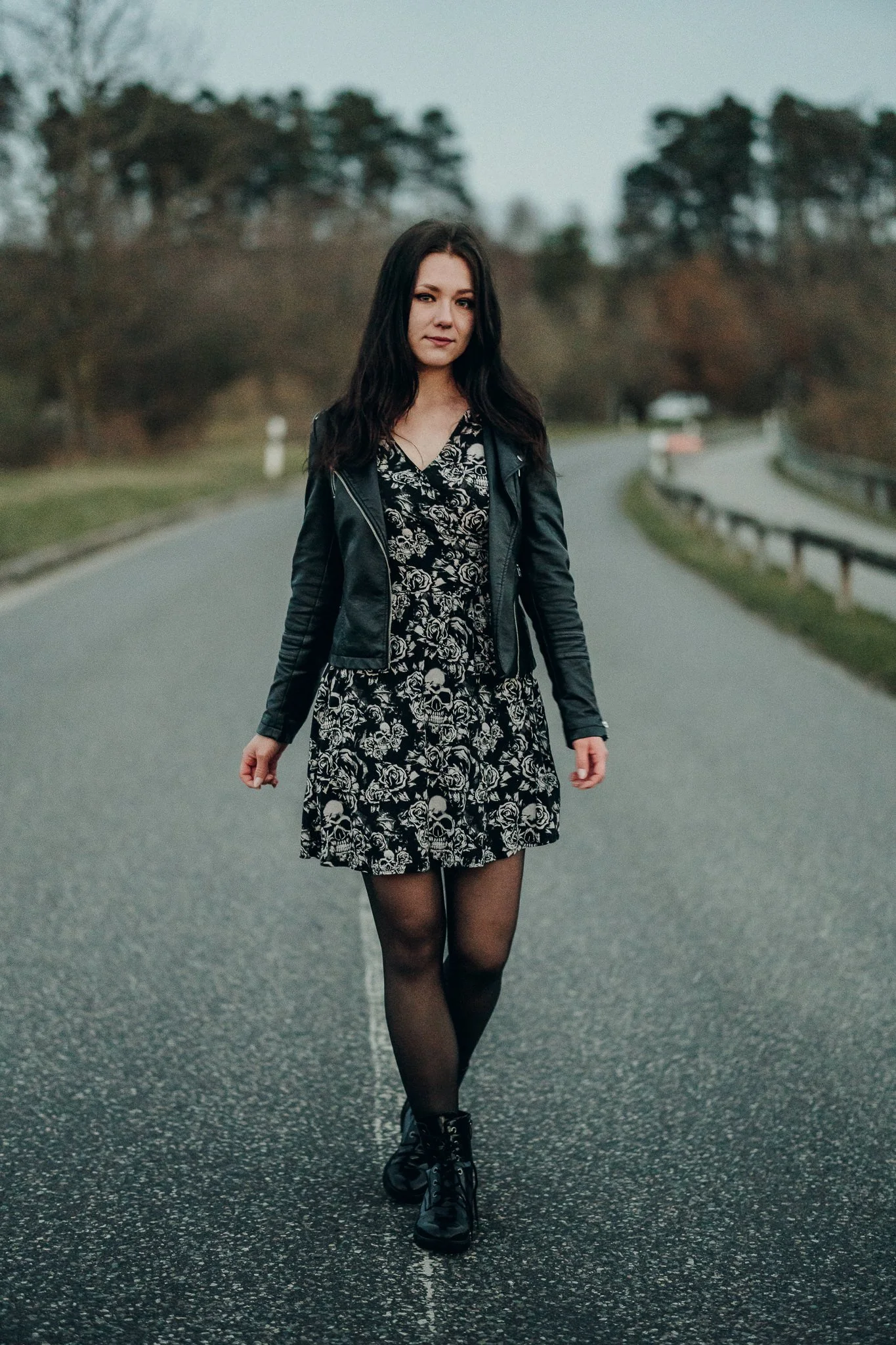 Eine junge Frau läuft auf einer leeren Straße in einer natürlichen Umgebung bei Dämmerung, trägt ein schwarzes Kleid mit Totenkopf- und Rosenmuster, schwarze Stiefel und eine schwarze Lederjacke.
