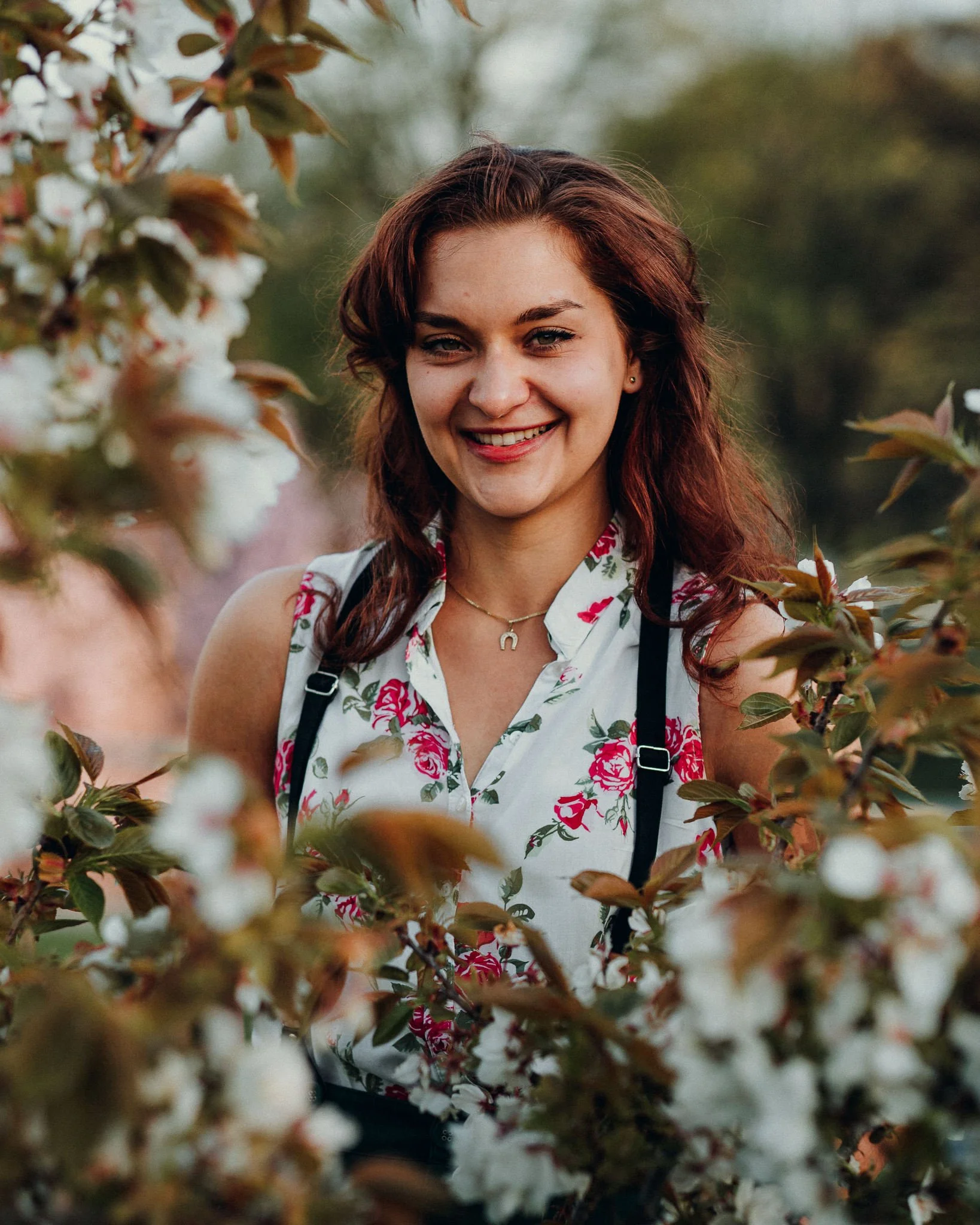Eine lachende junge Frau mit braunen Haaren und einem Blumenmuster-Hemd steht zwischen blühenden Zweigen. Es ist eine Outdoor-Aufnahme bei Sonnenuntergang oder Dämmerung.