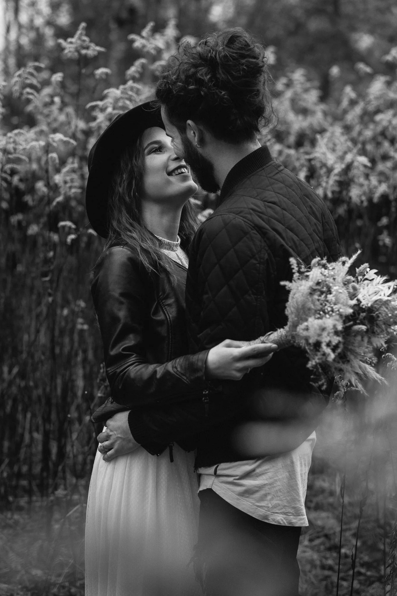 Paare, die sich im Freien bei Sonnenuntergang umarmen, eine Frau mit Hut und langer Rock, ein Mann mit Bart und Quiff, hält einen Blumenstrauß.