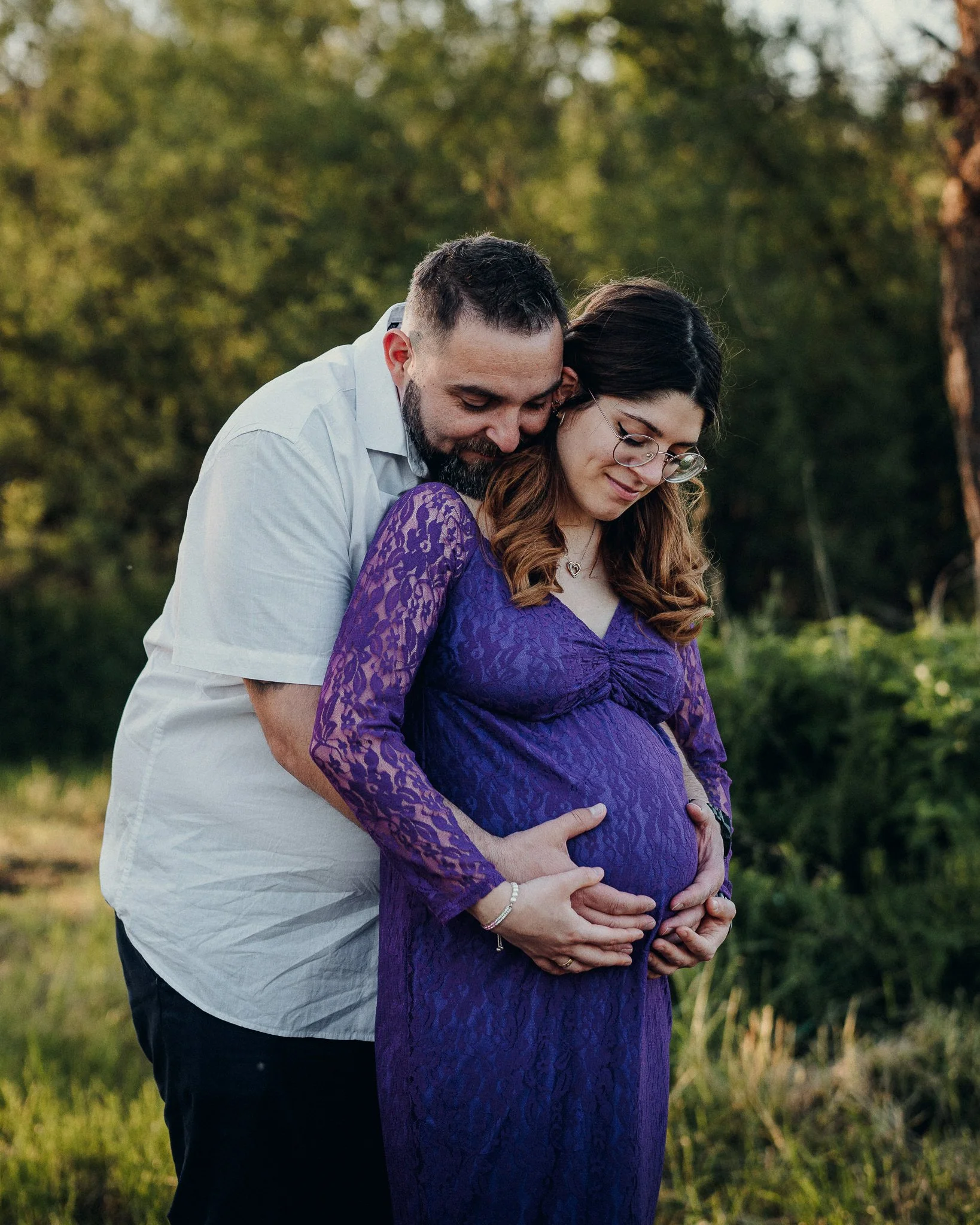 Ein schwangeres Paar, umarmt sich im Freien bei Sonnenuntergang, sie trägt ein lila Spitzenkleid, er trägt ein weißes Hemd.