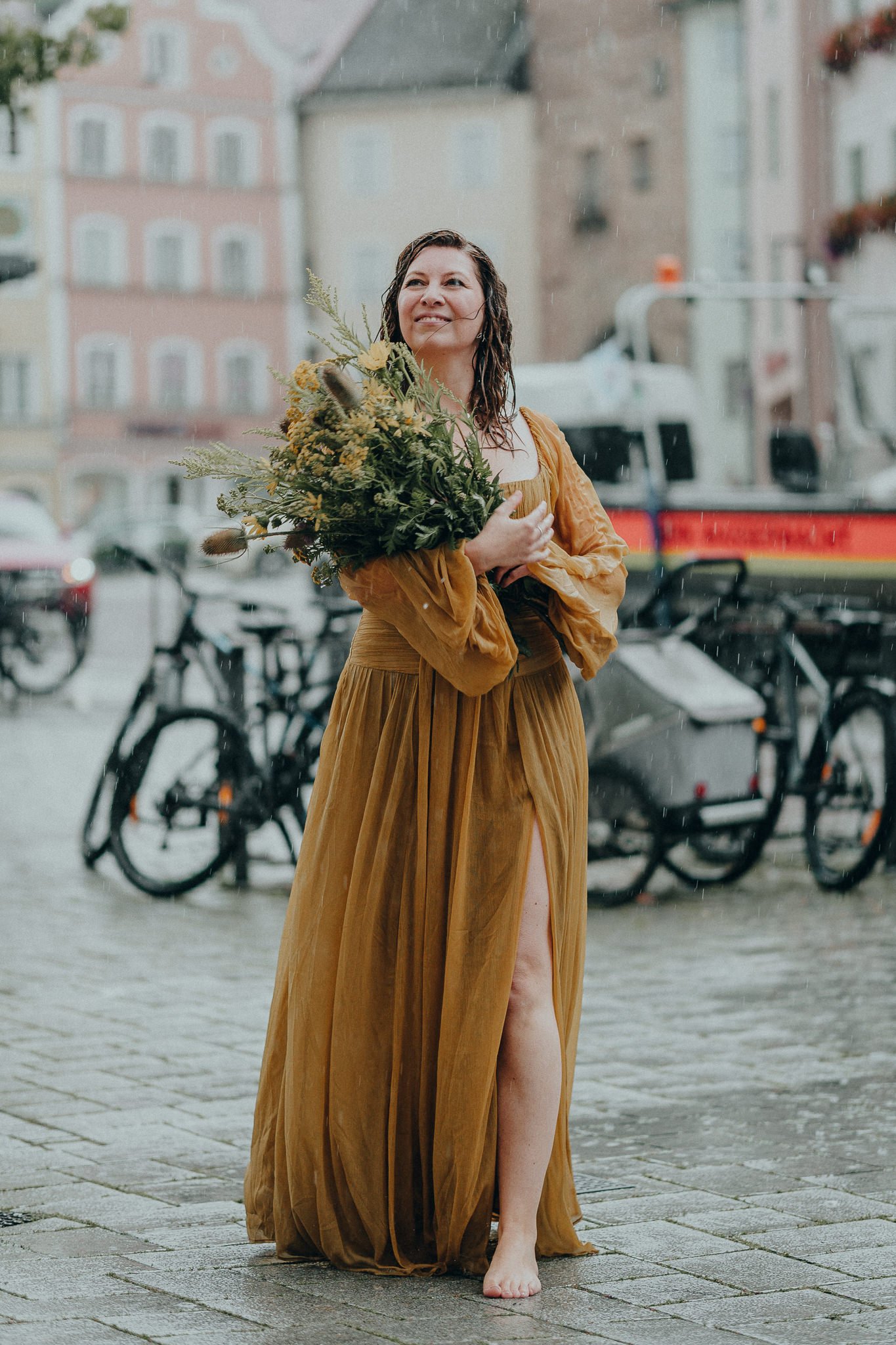 Eine Frau in einem langen, braunen Kleid steht barfuß auf dem Kopfsteinpflaster, hält einen Blumenstrauß und lächelt bei Regen vor einem historischen Stadthintergrund.