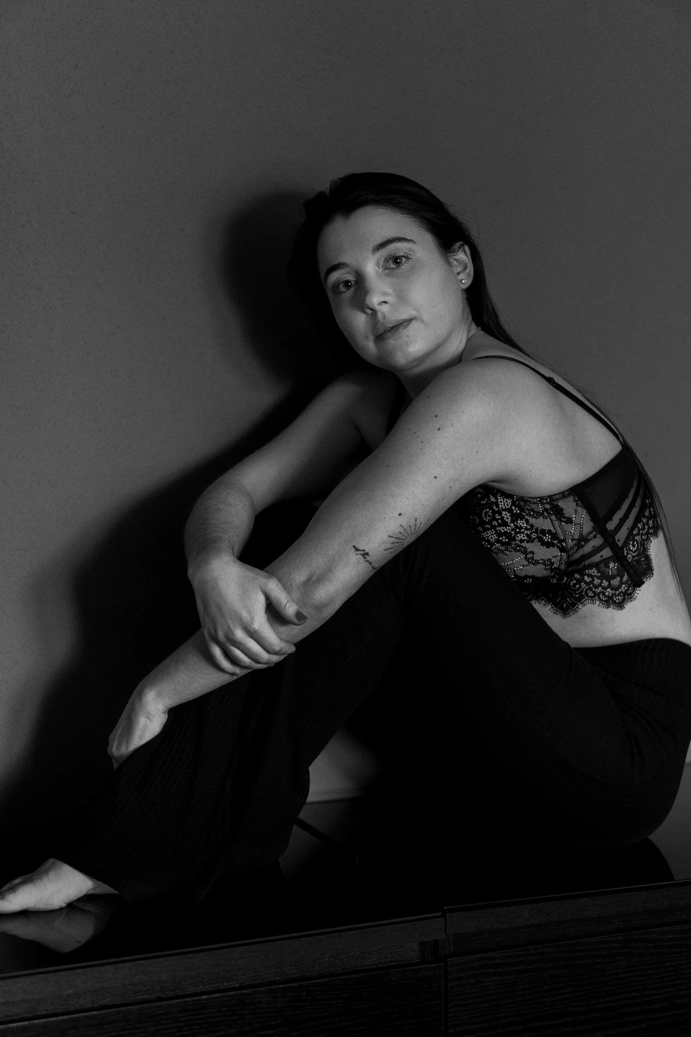 Schwarz-weiß-Foto einer jungen Frau, die in einem schwarzen Spitzenoberteil sitzt, mit einem Arm um das Knie und dem Blick direkt in die Kamera.