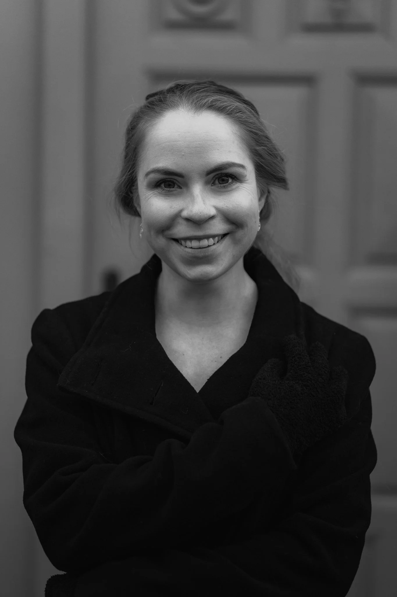 Schwarz-Weiß-Foto eines lachenden jungen Mädchens mit schulterlangem Haar, das eine Jacke trägt und vor einer Holztür steht.