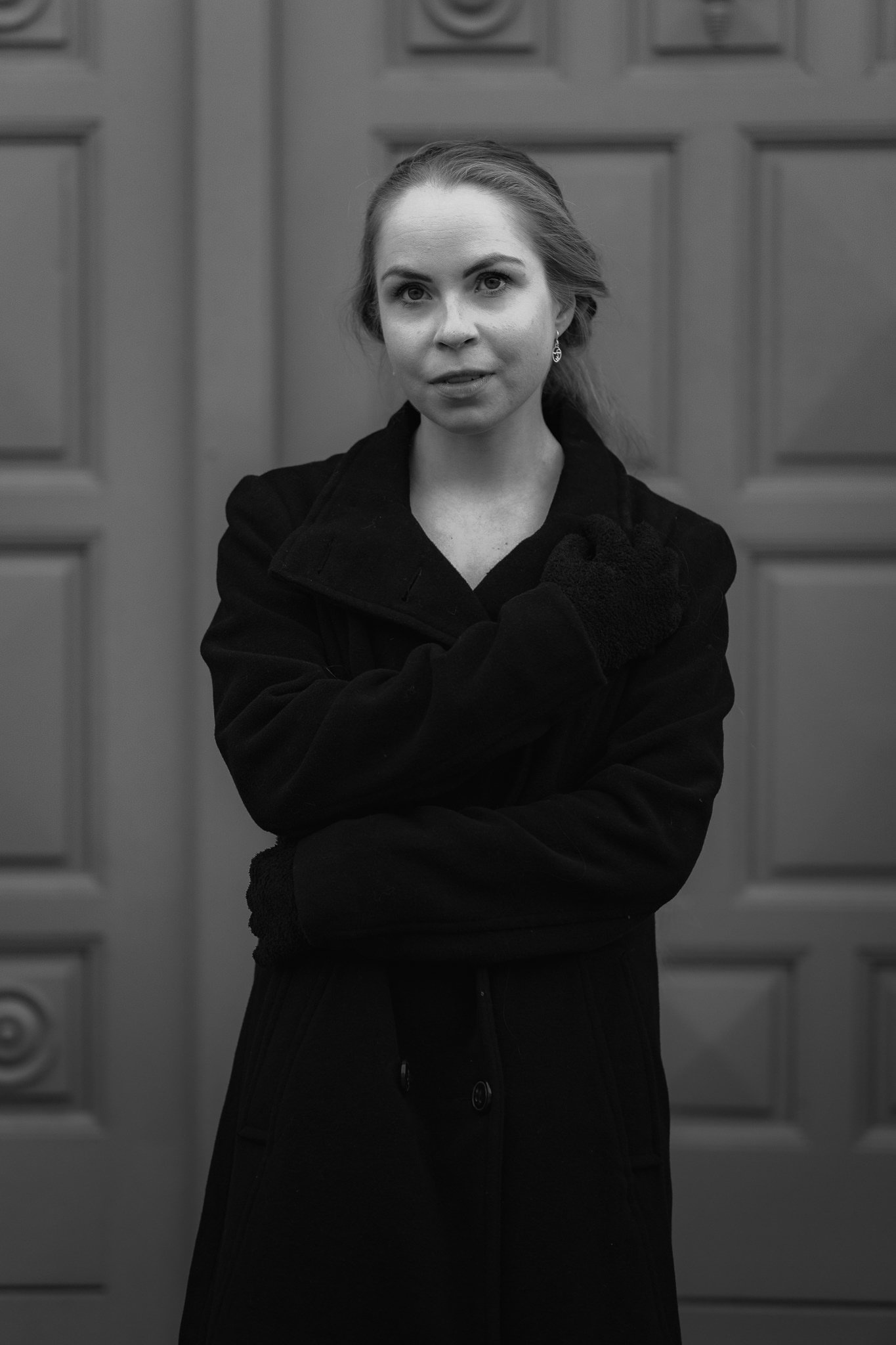 Schwarz-Weiß-Foto einer Frau in dunkler Kleidung vor einer Holztür, mit lockerer Frisur und einer Hand am Hals